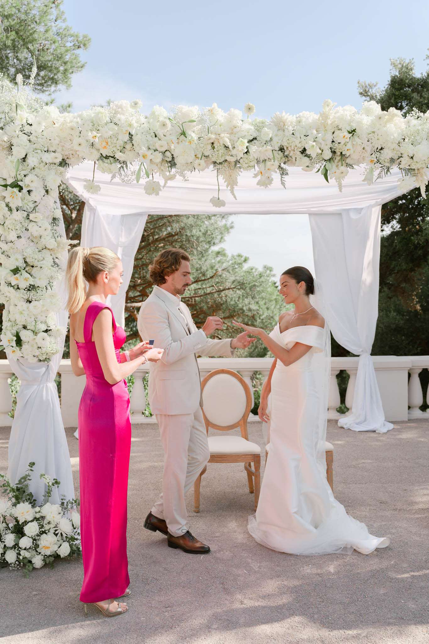 Ring exchange under white draped canopy with all-white floral arch on terrace with balustrade railings