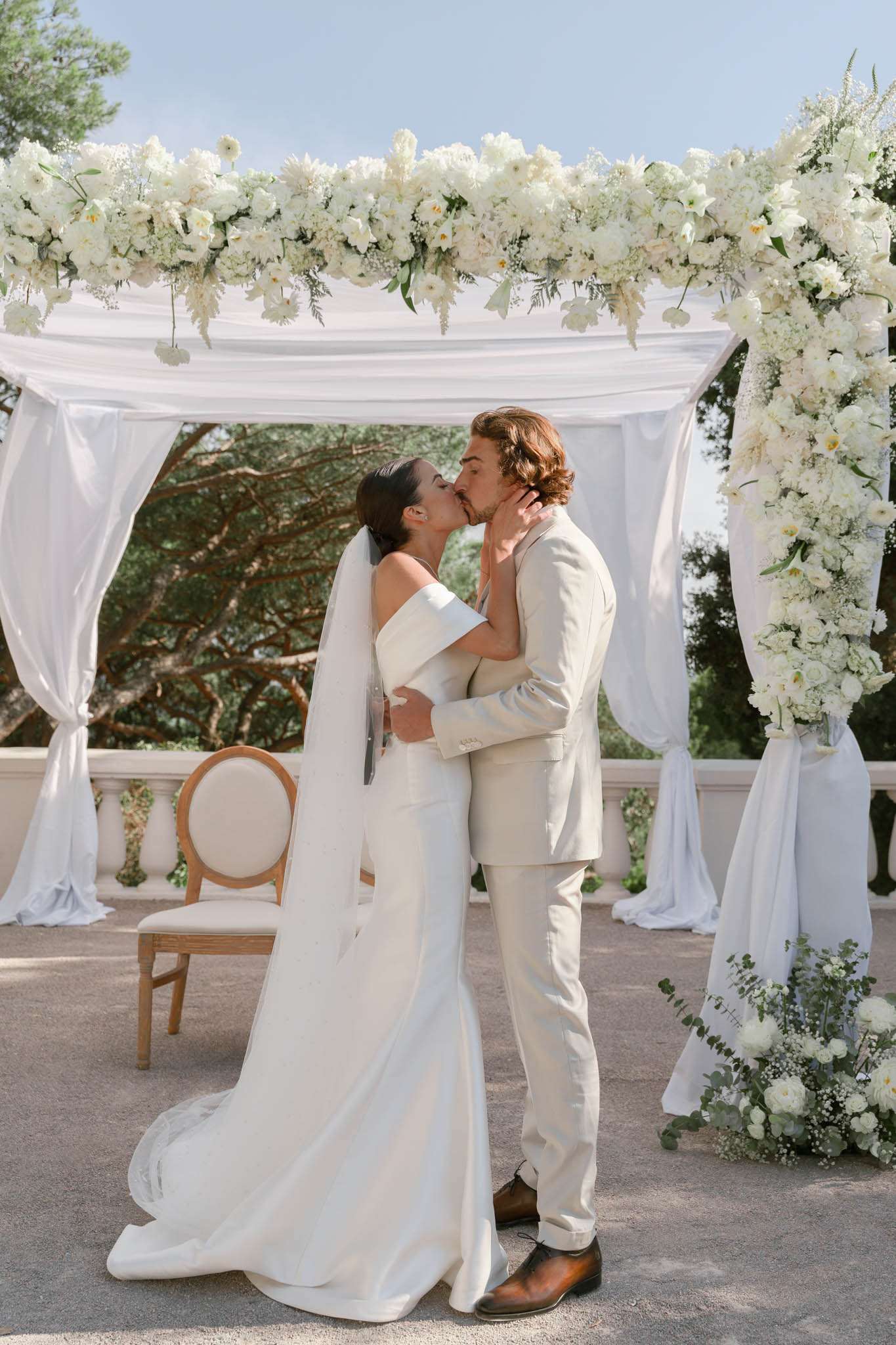 First kiss under white rose and wisteria draped arch, bride in cathedral veil, groom in beige suit