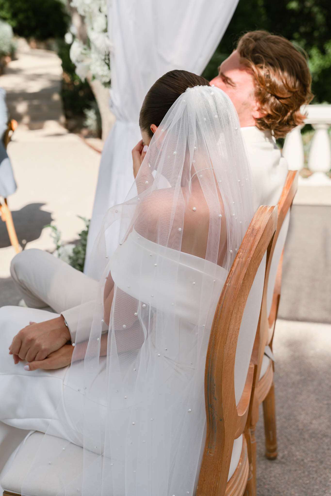 Bride and groom seated together during outdoor ceremony, photographed from behind showing pearl-embellished veil
