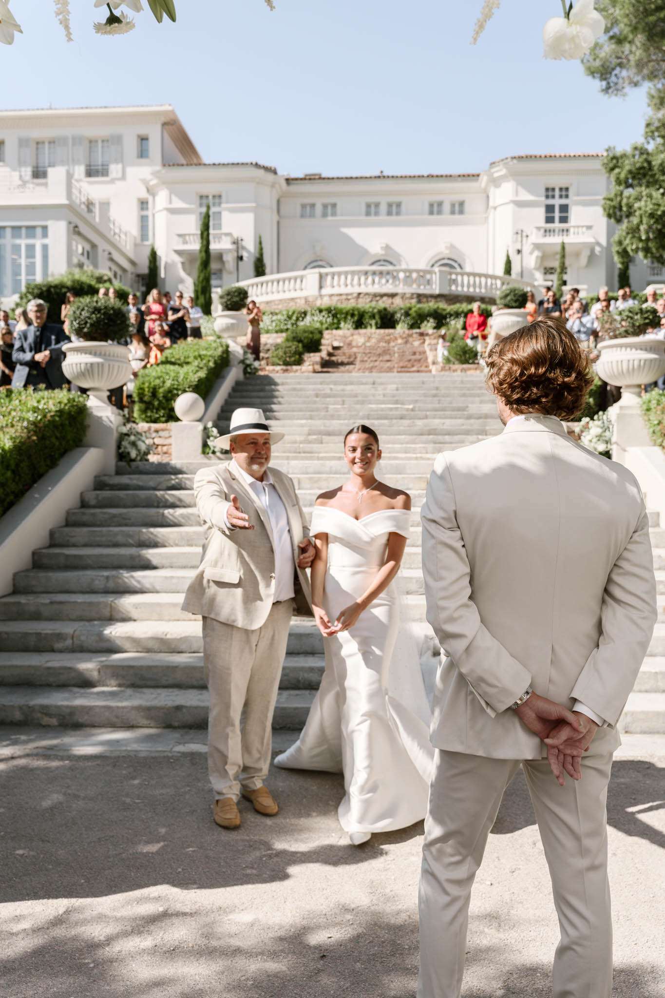 Bride escorted down grand stone staircase toward groom with seated guests and Mediterranean villa