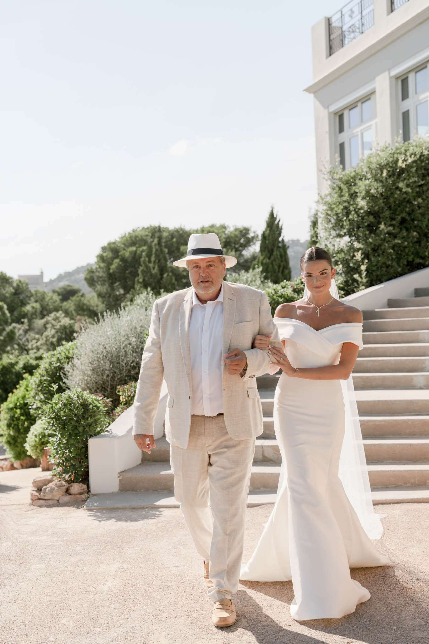 Bride in off-shoulder mermaid gown walking arm-in-arm with father in linen suit on villa terrace