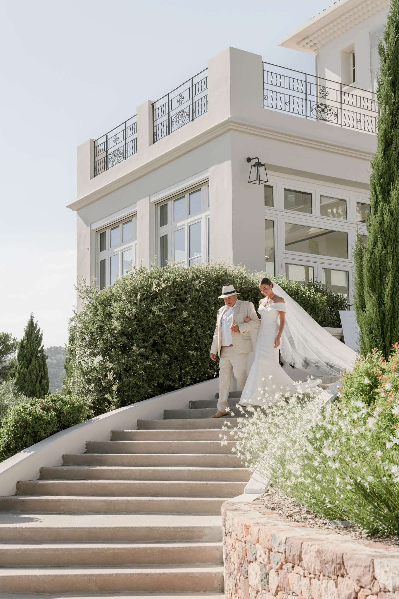 A bride and an older male companion — likely her father — walk arm-in-arm down a wide stone staircase outside a white rendered villa with black wrought-iron balcony railings and large French windows. The bride wears an off-the-shoulder ivory fitted gown with a long cathedral veil that billows in the wind behind her; her companion is dressed in a light beige suit with a white fedora hat. The setting is an outdoor terraced approach to what appears to be a South of France villa or château, with cypress trees framing the building and small white flowering plants in the foreground. The shot is a full-length wide portrait taken from the base of the stairs, capturing both figures and the full facade of the venue. Potential venue feature image.