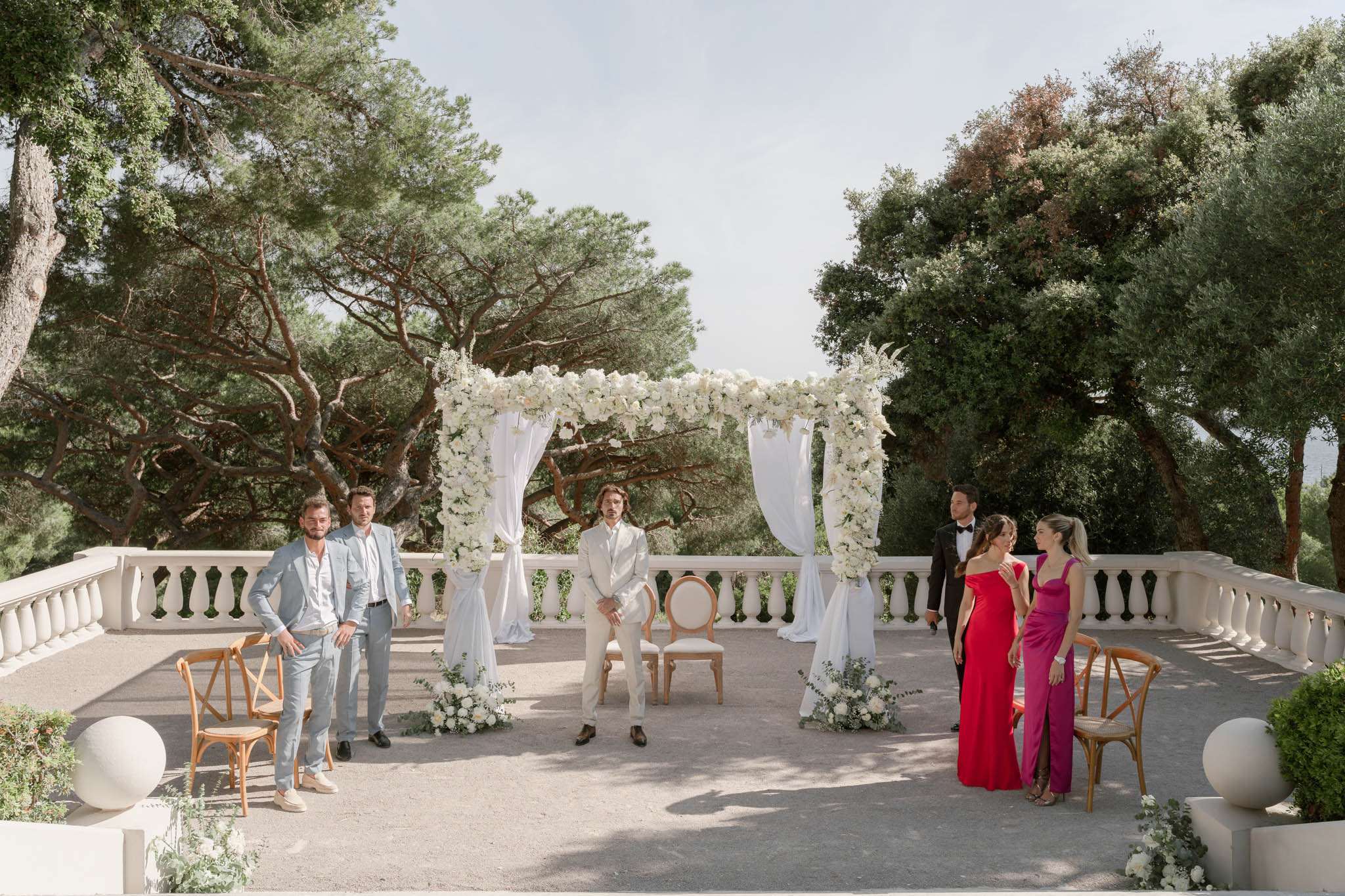 An outdoor ceremony setup on a white stone terrace with a classical balustrade, likely at a French villa or château. A rectangular arch draped with white fabric and densely covered in white roses and white blooms with trailing greenery serves as the ceremony focal point, flanked by white floral ground arrangements. A man in a cream suit is seated in a Louis XVI-style chair at the altar, appearing to be the groom waiting for the ceremony to begin. Two men in light blue and grey suits stand to the left, while on the right a man in a black tuxedo stands alongside two women in floor-length gowns — one in red and one in magenta pink. Wooden cross-back chairs are positioned on both sides of the terrace. The overall decor palette is white, ivory, and soft green, with a classic French style. Wide shot capturing the full ceremony space and wedding party.