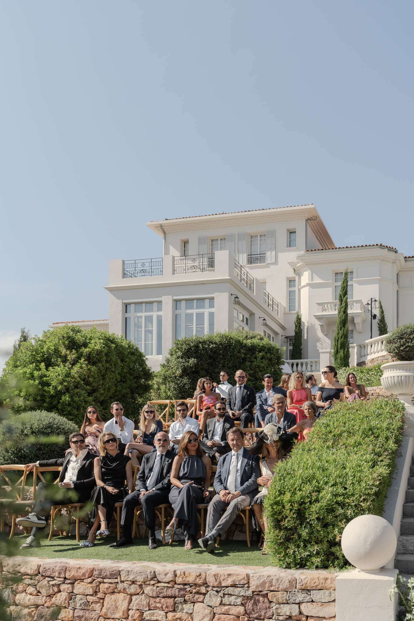 30 guests on tiered lawn with crossback chairs facing white multi-storey villa with cypress trees