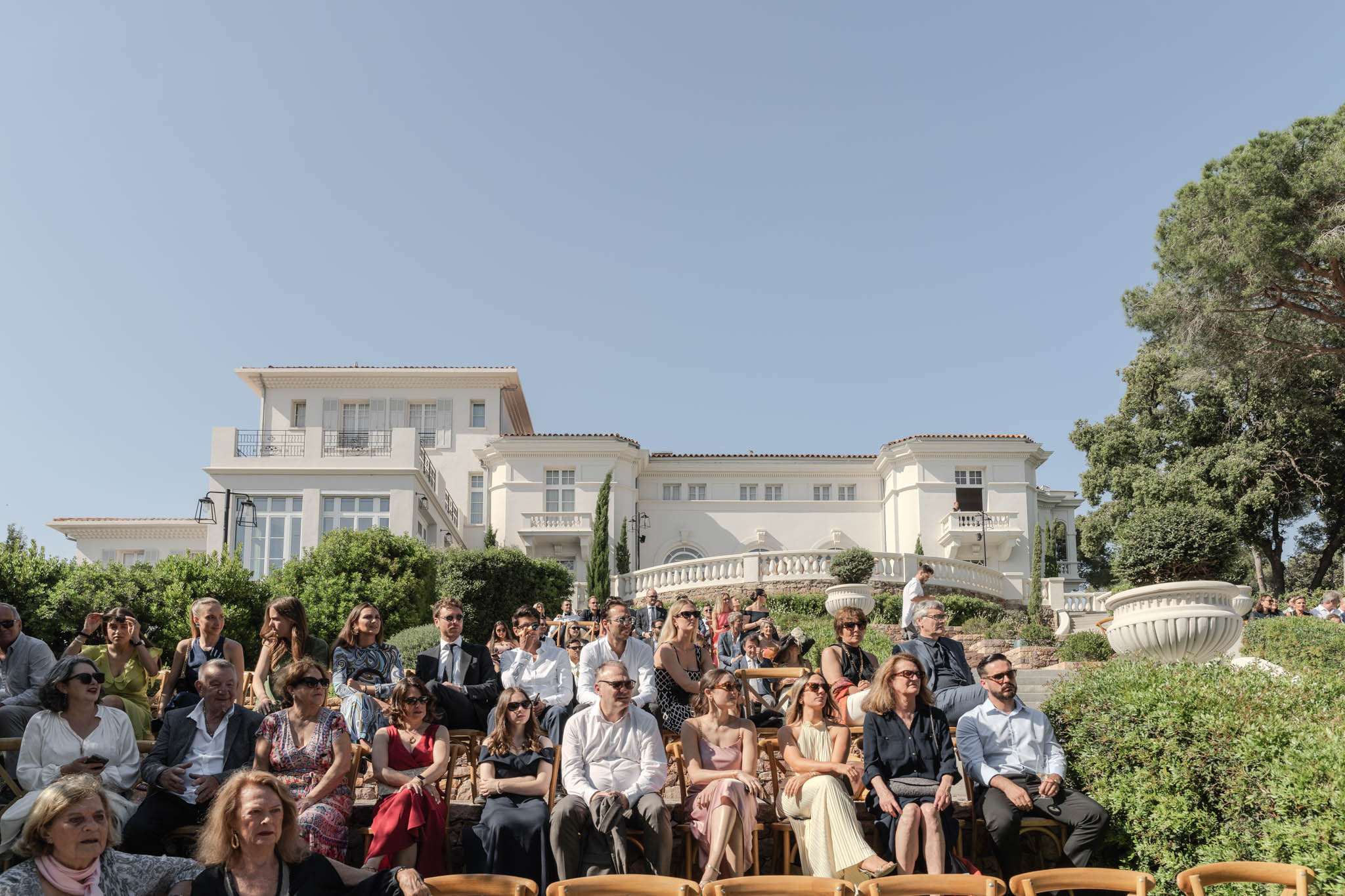 An outdoor wedding ceremony is in progress, with approximately 40–50 guests seated on wooden folding chairs arranged in rows facing the officiant or couple (out of frame). The setting is the terraced grounds of a large white Mediterranean-style villa with terracotta roof tiles, wrought-iron balconies, and a classical balustrade terrace visible directly behind the guests. Guests are dressed in smart-casual to semi-formal attire in a mix of whites, blacks, reds, and soft pinks, with many wearing sunglasses suggesting a bright, warm daytime event. The wide-angle shot is taken from a low angle facing the crowd, placing the white villa prominently in the background. Potential venue feature image.