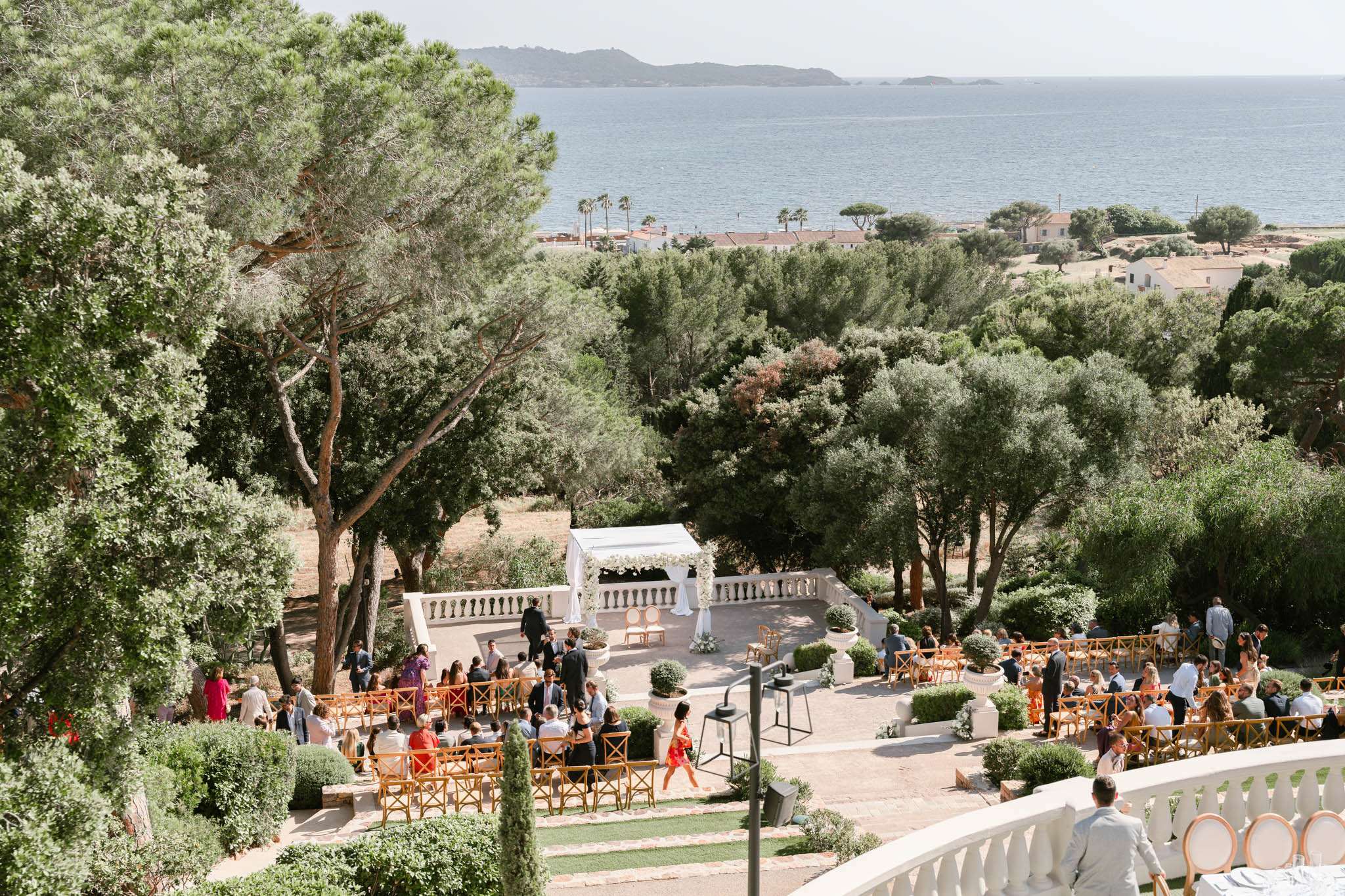 Elevated view of seaside terrace ceremony with white floral arch cross-back chairs and 80 guests