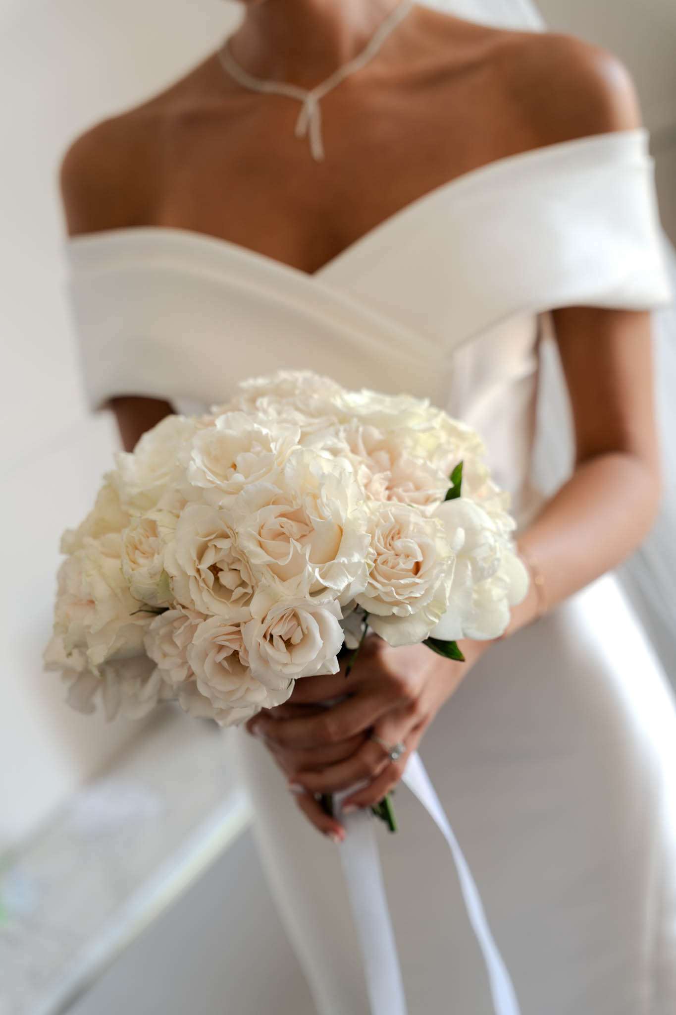 Close-up of bride in off-shoulder satin gown holding round bouquet of blush garden roses and white peonies