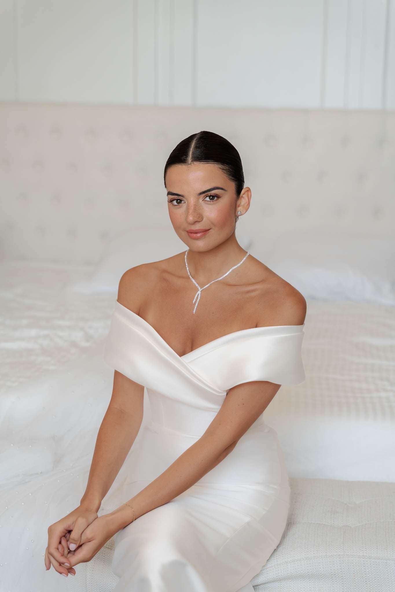 Bride seated on white bed wearing ivory off-the-shoulder satin gown with diamond necklace and sleek bun