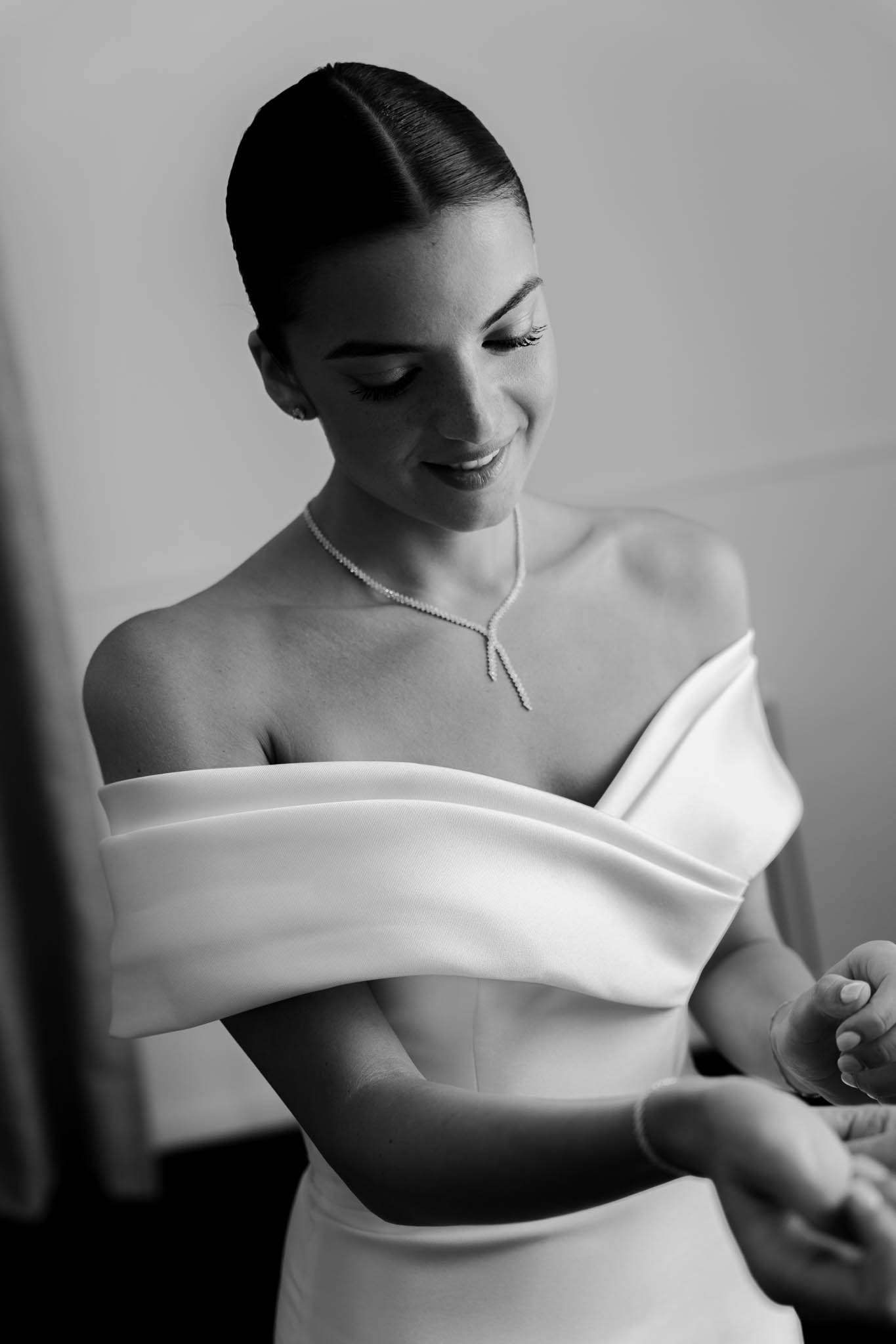 Bride smiling as assistant fastens detail on off-shoulder gown with structured folded bodice in B&W
