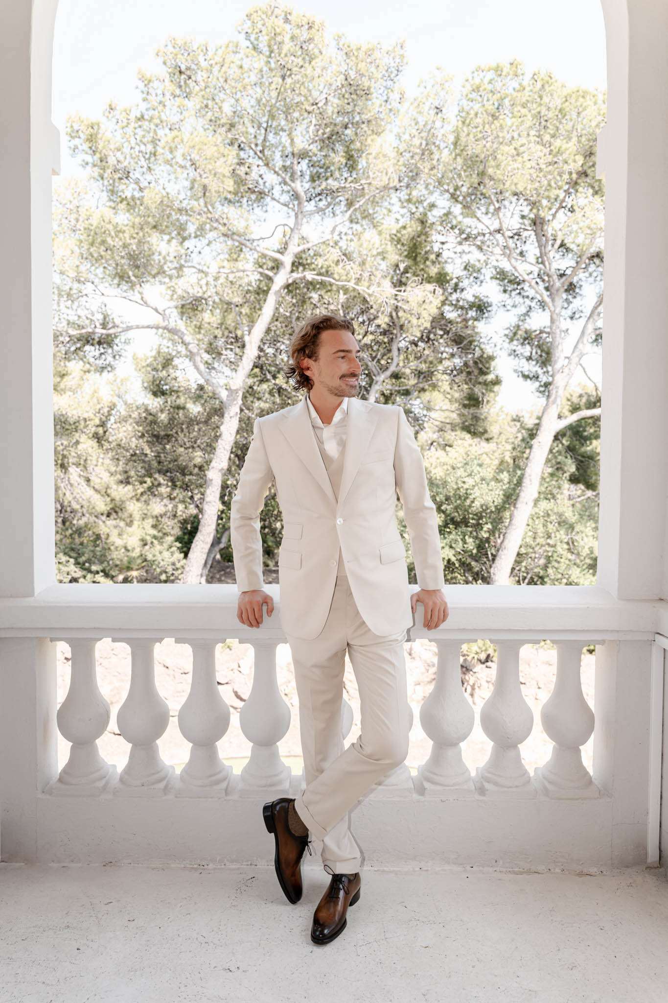 Groom in cream three-piece suit leaning against white stone balustrade terrace with trees behind