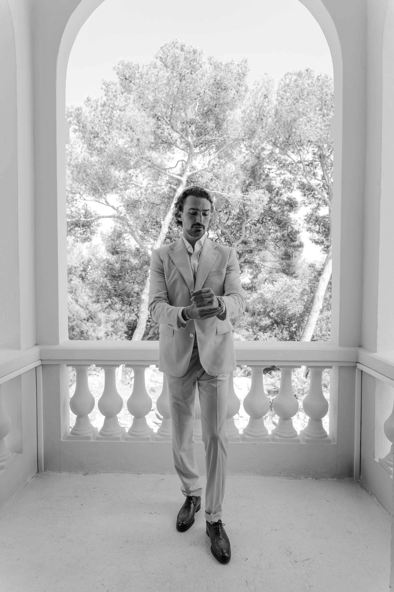 Black-and-white portrait of groom adjusting cufflinks on chateau terrace with arched opening and pine trees