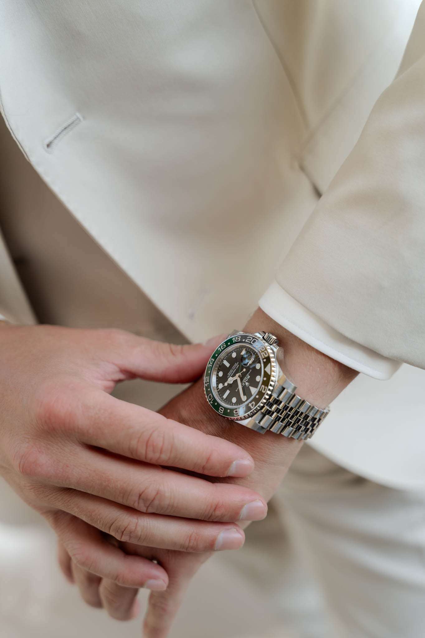 A close-up detail shot of a groom's wrists during the getting-ready phase, showing one hand adjusting a stainless steel sport watch with a green bezel and black dial on the other wrist. The groom is wearing an ivory or cream suit jacket. The composition is a tight portrait-style detail crop focusing on the hands and watch as a styling accessory.