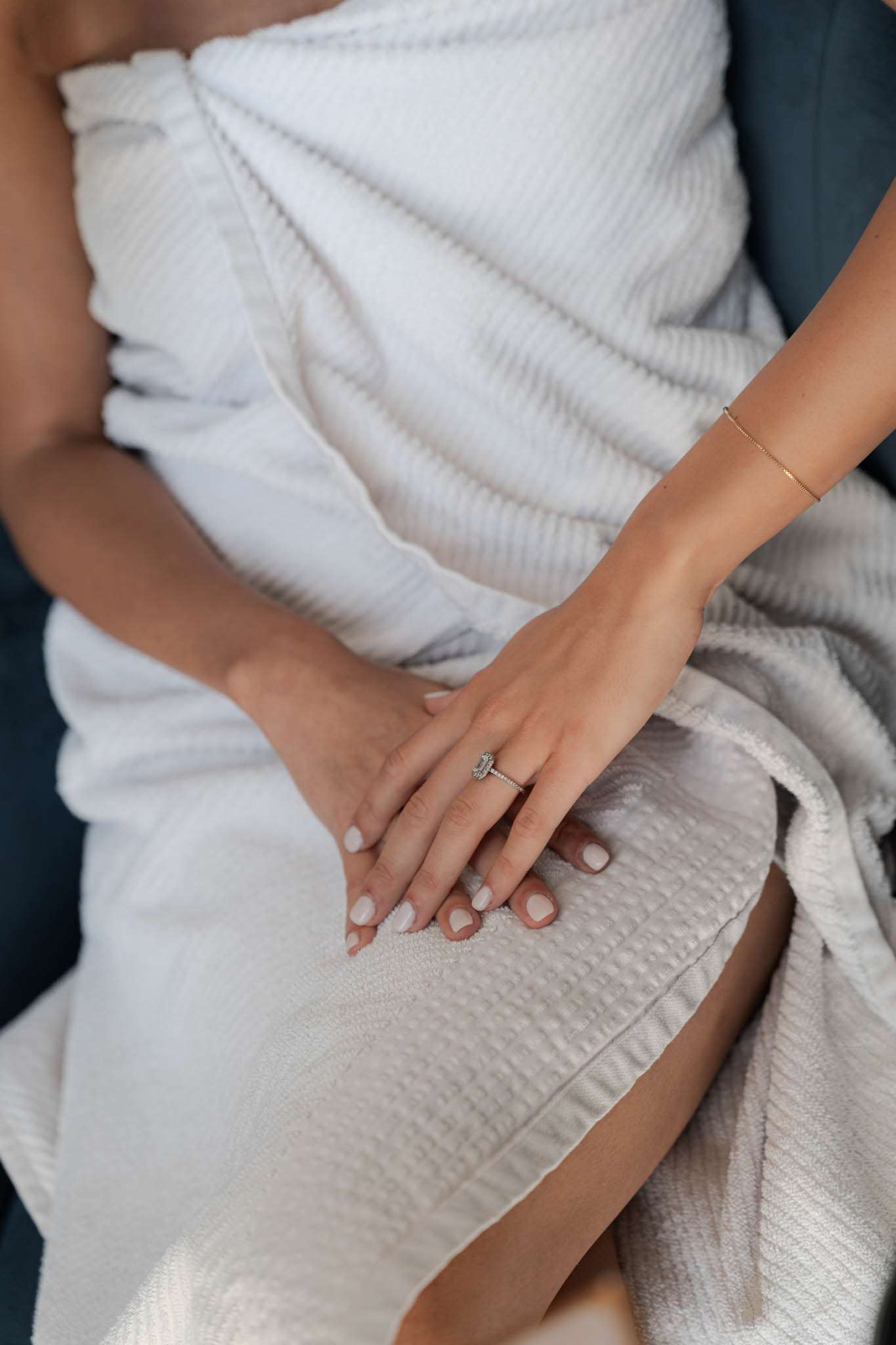 Close-up bride's hands in white waffle robe showing marquise diamond ring and gold chain bracelet