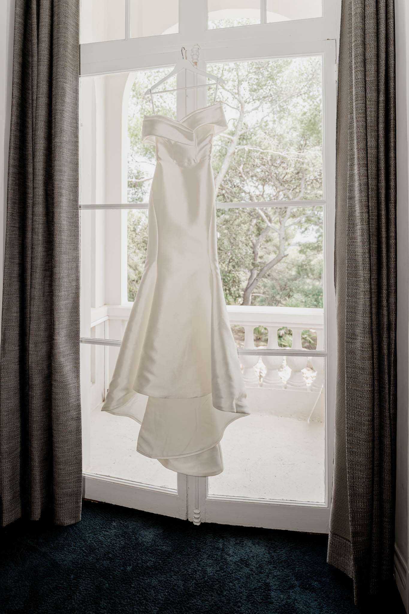 Ivory satin off-the-shoulder wedding dress hanging on a clear hanger in front of tall white French doors