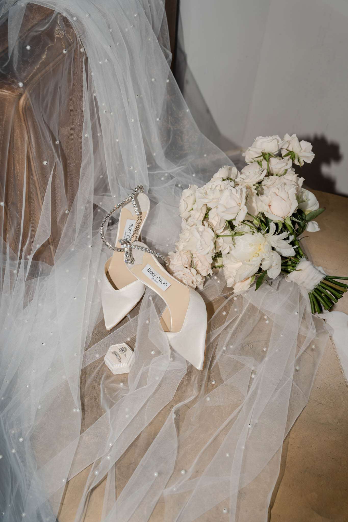 A bridal detail flat lay arranged on a upholstered surface, featuring a pearl-scattered tulle veil draped across the scene. A pair of ivory satin Jimmy Choo pointed-toe heels with crystal embellished ankle straps are placed at the center, alongside a bouquet of blush and white garden roses, peonies, and white freesia with green stems wrapped in white ribbon. A small white hexagonal ring box containing what appears to be a diamond engagement ring and wedding band is tucked near the base of the shoes. The overall color palette is ivory, blush, and crystal white with a classic, polished styling approach. Close-up detail shot with natural indoor lighting.