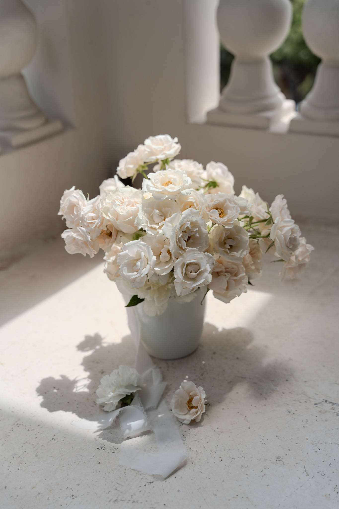 A floral detail shot featuring a compact arrangement of cream and blush garden roses in a white ceramic vessel, placed on a pale stone ledge or balustrade. Two loose rose heads and a short length of white satin ribbon are scattered on the surface beside the vase. The background shows white classical stone balusters, suggesting an outdoor terrace or formal garden setting. Natural sunlight casts sharp shadows across the pale stone surface, contributing to a bright, all-white and cream color palette with a classic, refined styling approach.