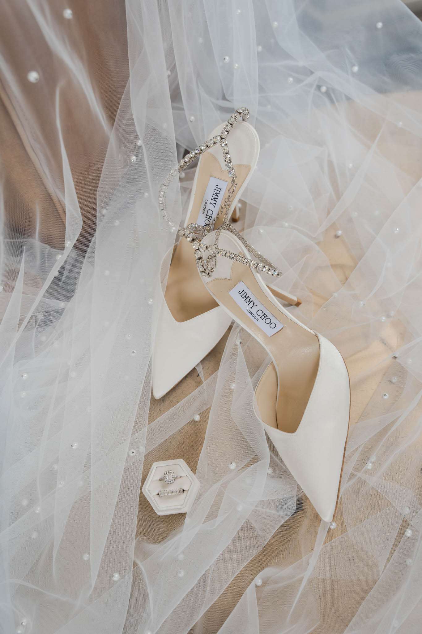 A close-up flat lay of bridal accessories arranged on a pearl-embellished tulle veil. The centerpiece is a pair of ivory Jimmy Choo pointed-toe heels with crystal-encrusted ankle straps and toe straps. In the lower left corner, a small hexagonal ivory velvet ring box holds two rings — an emerald-cut diamond engagement ring and a diamond wedding band. The pearl detailing on the veil is scattered throughout the composition, reinforcing a classic bridal aesthetic. The overall color palette is ivory and white with silver crystal accents.