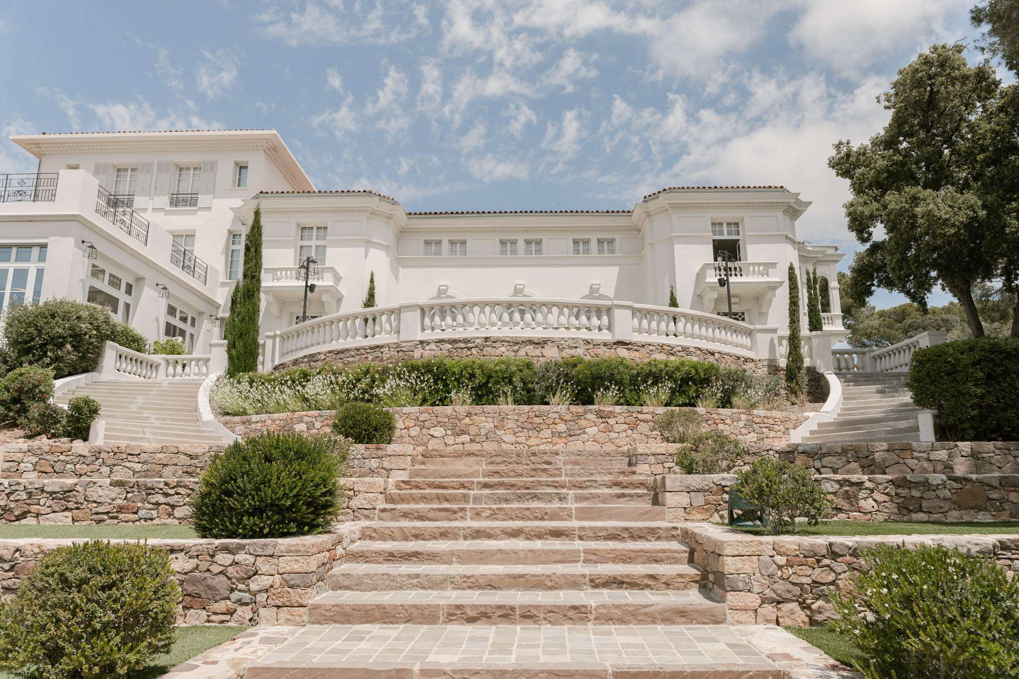 An exterior wide shot of a large white villa-style venue featuring multi-level terraced grounds connected by broad sandstone steps leading up from a paved pathway. The architecture is classical Mediterranean in style, with white rendered facades, terracotta-tiled rooflines, wrought iron balconies, and a curved white balustrade terrace at the upper level flanked by tall cypress trees. The terracing incorporates dry-stone retaining walls in warm sandy tones, with manicured round-clipped shrubs and low flowering plants placed throughout the landscaped levels. No people are visible in this image. Potential venue feature image.
