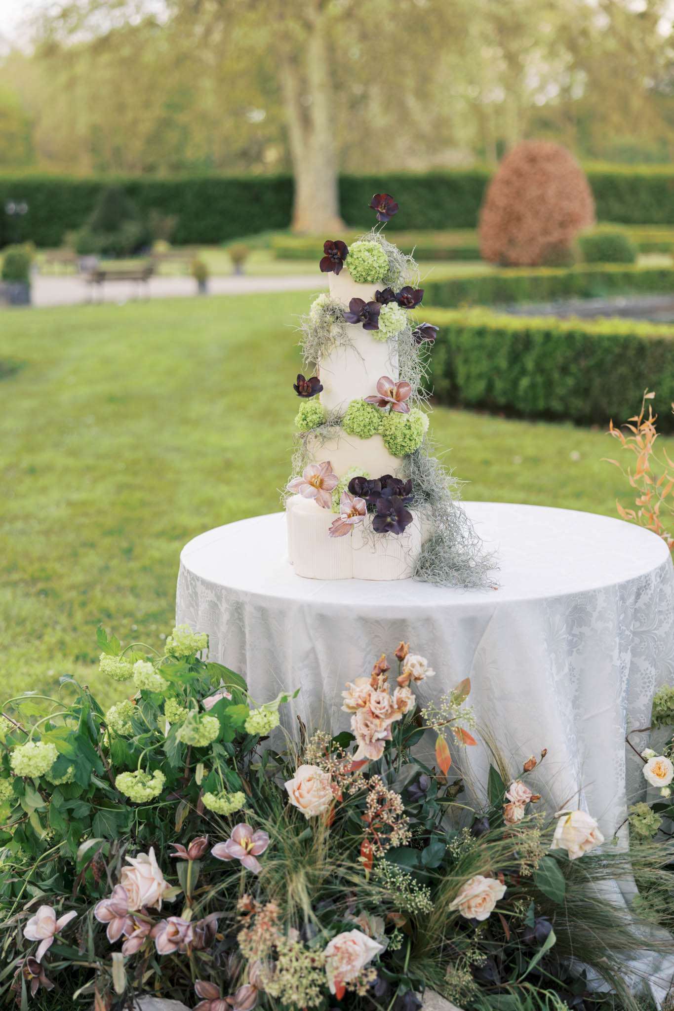 Five-tier white cake with cascading burgundy orchids, blush roses, and trailing Spanish moss in garden