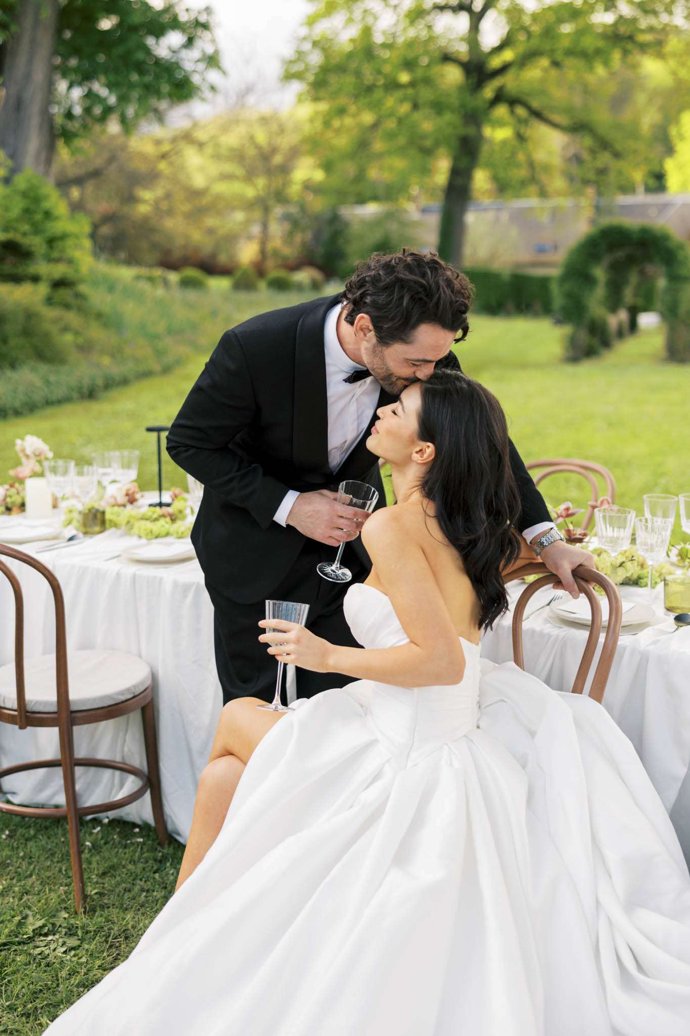 Groom kissing seated bride on forehead at outdoor reception table with greenery arrangements and crystal glassware