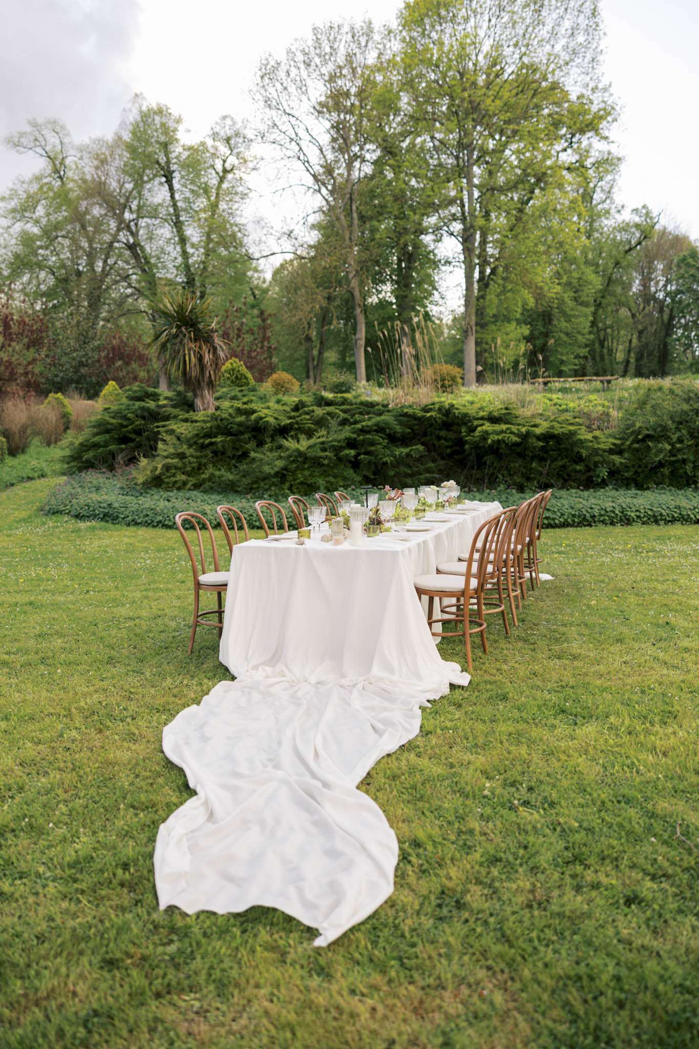 An outdoor wedding reception table is set on a garden lawn, styled for an intimate gathering of approximately 10–12 guests. The long rectangular table is covered with a white linen tablecloth that dramatically pools and trails along the grass toward the foreground, a deliberate styling choice. Bentwood chairs with warm honey-toned wood frames and cream upholstered seats are arranged along both sides of the table. The tablescape features glassware, white plates, small floral arrangements with soft pink blooms and greenery, and what appear to be bud vases and candle vessels centered along the table. The overall decor palette is white and natural wood with soft pink floral accents, reflecting a classic garden aesthetic. Wide shot taken from ground level, emphasizing the trailing tablecloth as a focal compositional element.