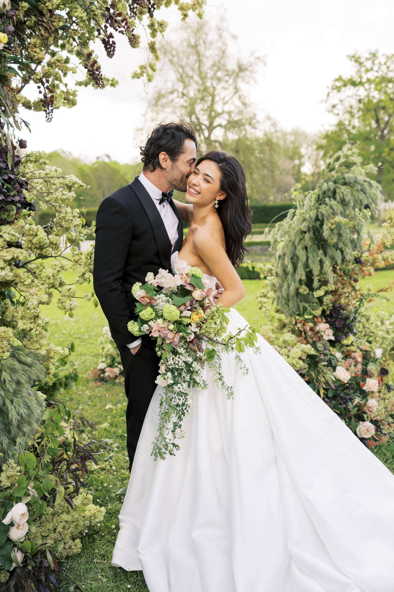 An outdoor couple portrait taken in a formal garden setting, showing the bride and groom standing within a large floral arch or installation. The groom, wearing a black tuxedo with a navy bow tie, kisses the bride on the cheek while she laughs and faces the camera. The bride wears a strapless ivory ball gown with a full skirt and holds a large cascading bouquet composed of green viburnum snowball clusters, blush roses, dusty pink hellebores, yellow ranunculus, trailing greenery, and white filler florals. The floral arch surrounding them features the same green viburnum, deep burgundy blooms, blush garden roses, and trailing foliage in a lush, garden-style arrangement. The overall styling is romantic and garden-inspired with a soft palette of blush, ivory, green, and deep plum. The shot is a mid-length portrait framing the couple centrally against the floral backdrop and a manicured park landscape behind.