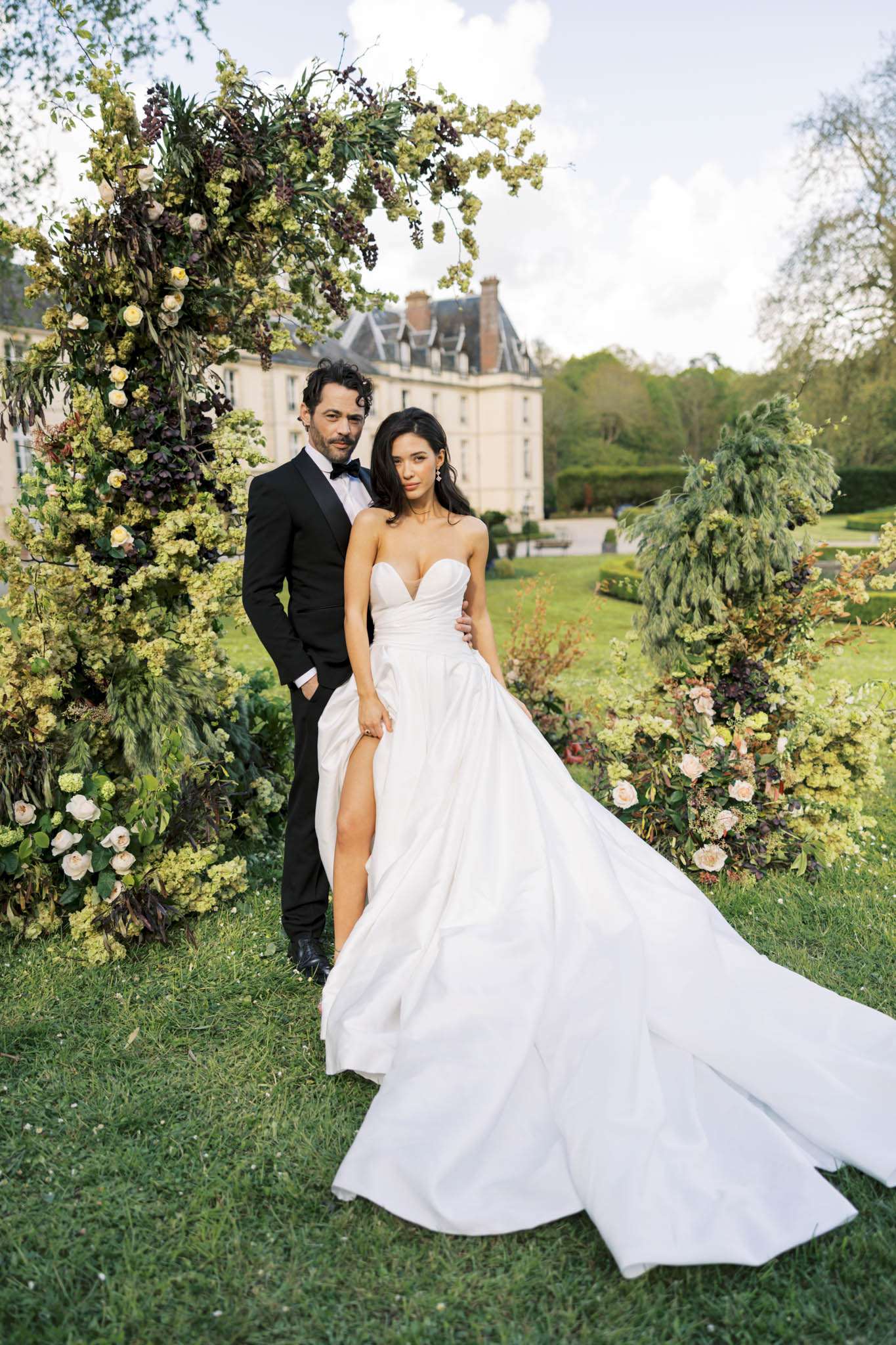 Bride in strapless ball gown and groom in tuxedo before a circular floral arch of hydrangeas and roses at a chateau