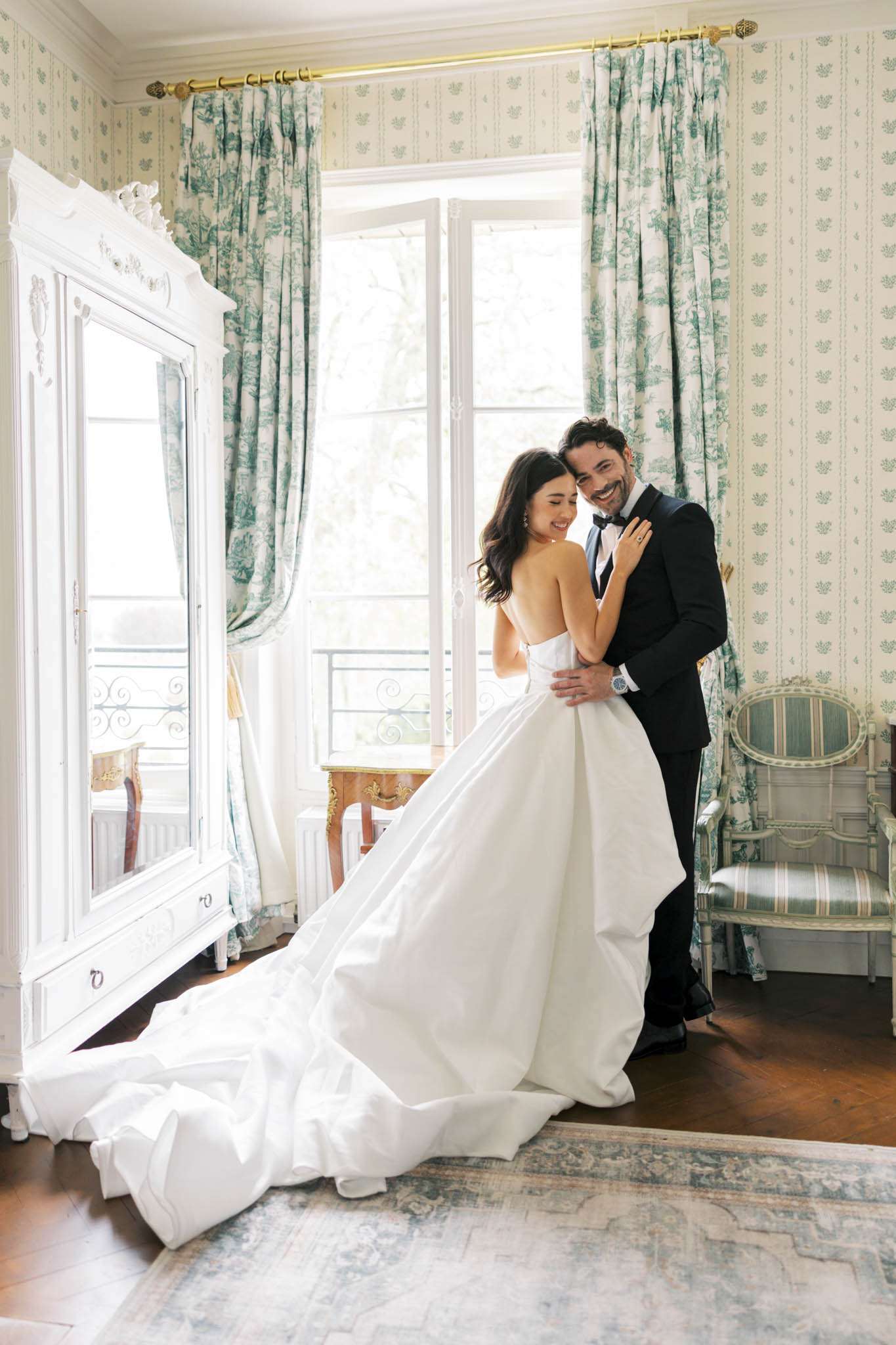 Bride in white ballgown and groom in black tuxedo laughing together in a toile de Jouy decorated chateau room