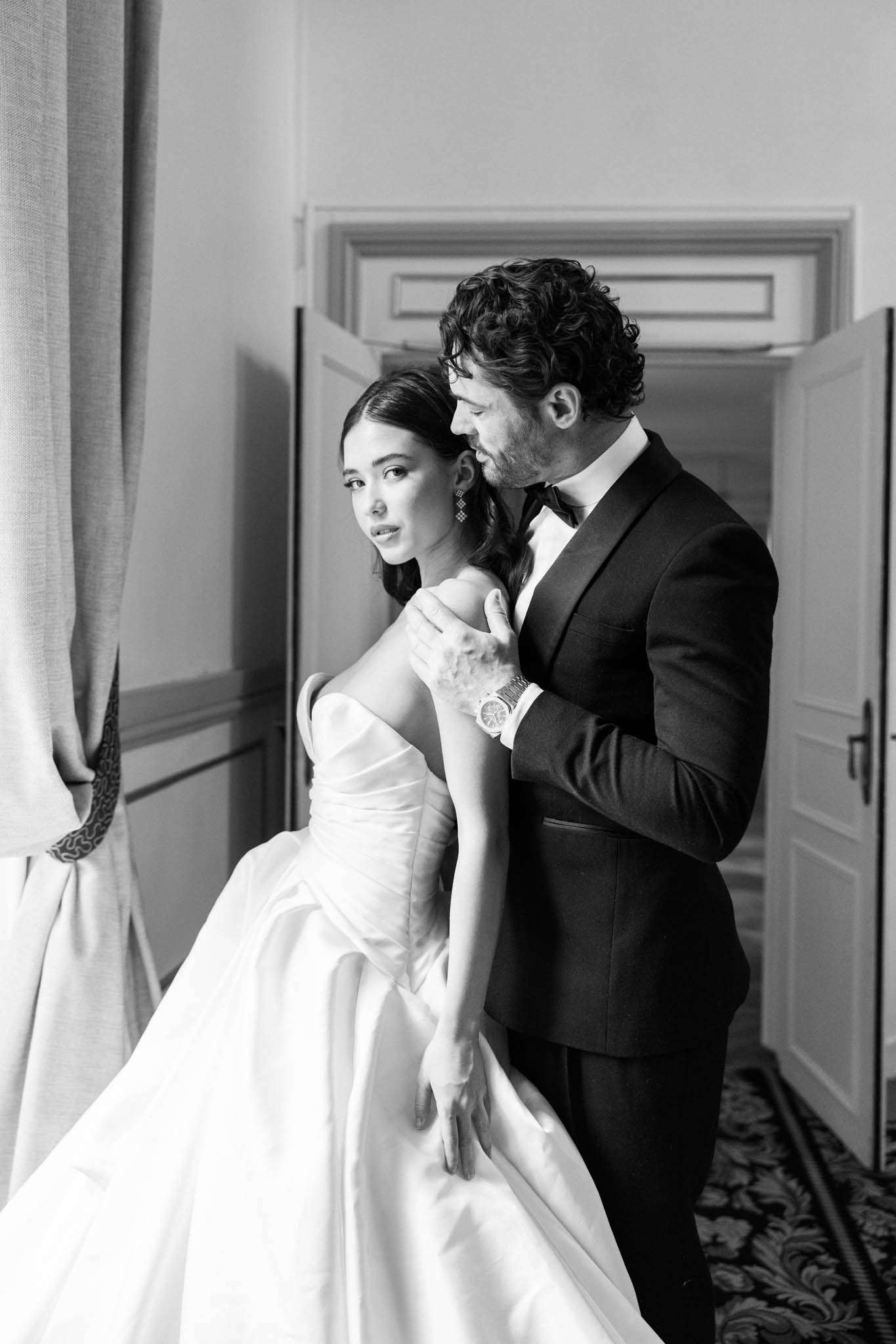 This is a black-and-white couple portrait taken indoors, likely inside a château or grand venue, with ornate molding above a set of double doors and a patterned carpet visible on the floor. The groom, wearing a dark tuxedo with a bow tie and a dress watch, stands behind the bride with his hands on her shoulders, leaning in close to her. The bride wears a strapless structured ballgown with a draped bodice and a full skirt, and she looks directly into the camera while wearing drop earrings. The image has strong contrast with bright highlights on the bride's dress and soft mid-tones on the interior walls and drapery. The composition is a close-to-medium portrait shot framed near a window with linen curtains to the left.