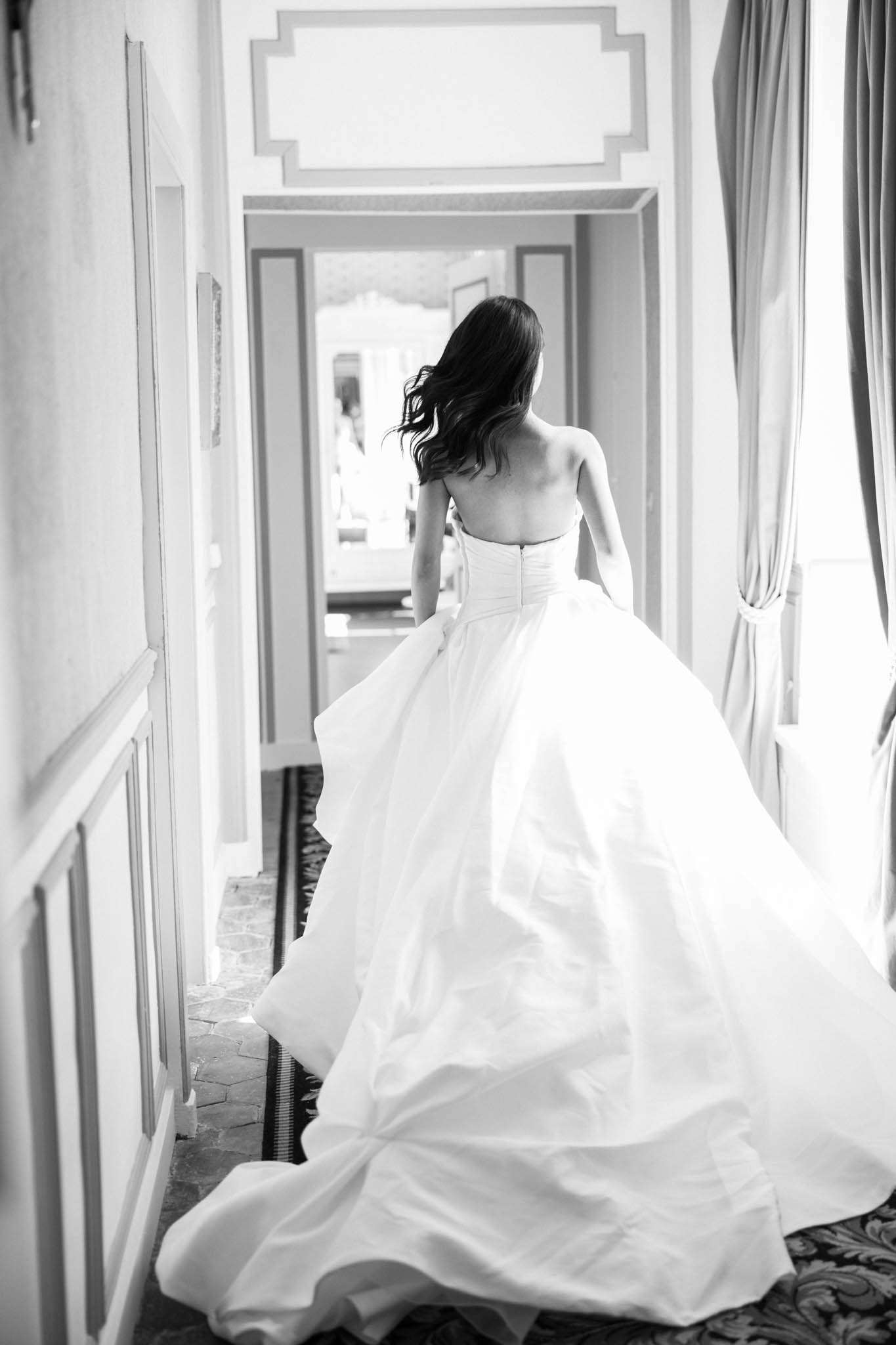 Black-and-white portrait of bride walking away down a chateau corridor in strapless ball gown with long train