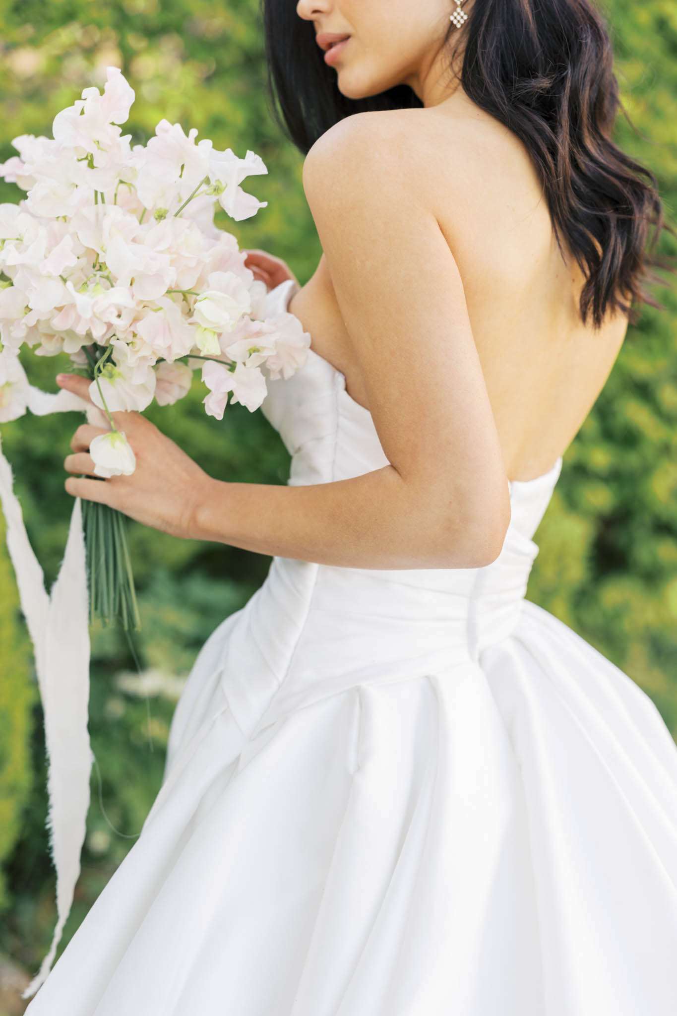 Bride from behind holding blush pink sweet pea bouquet with trailing silk ribbons, wearing strapless ballgown