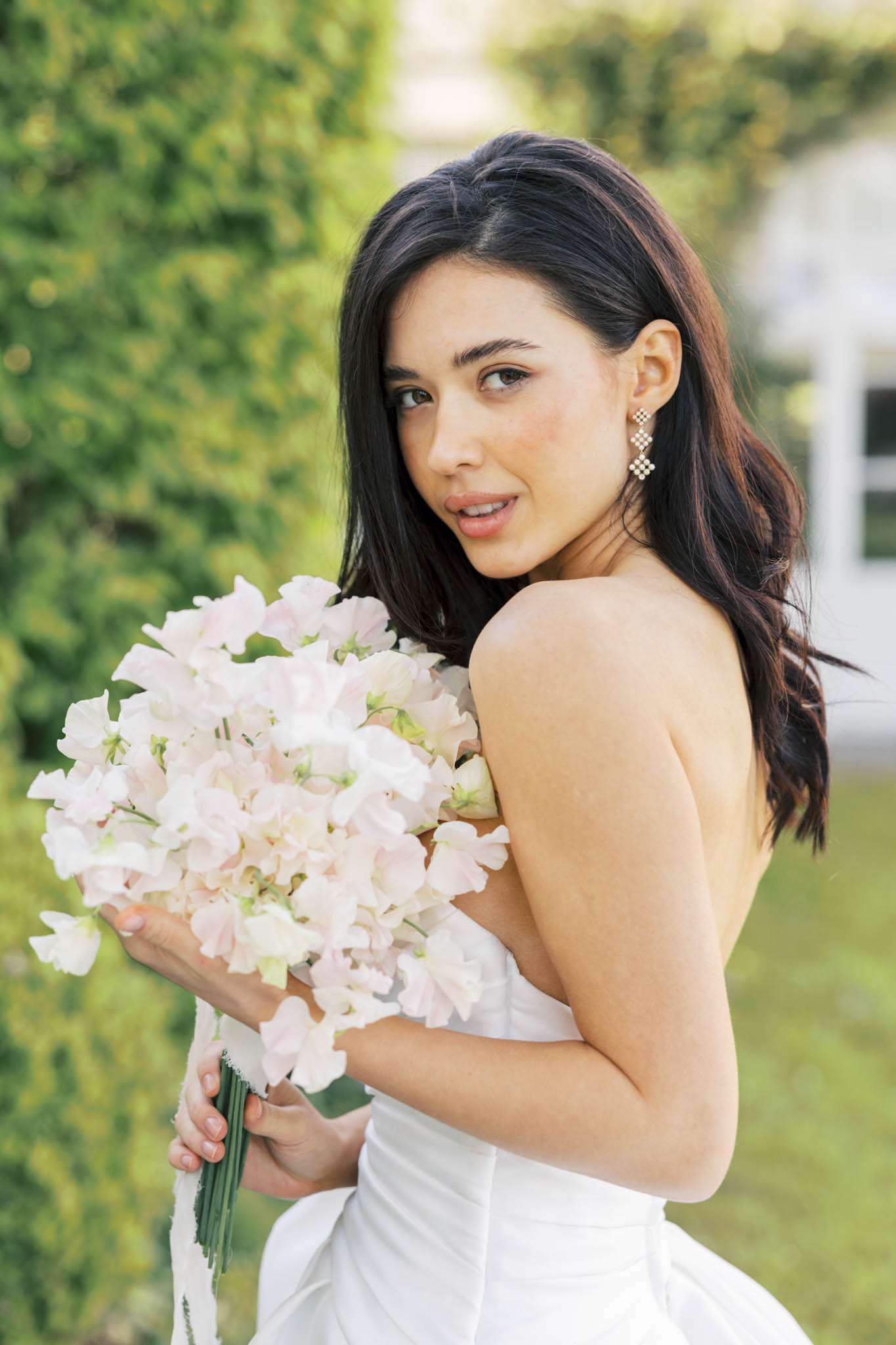 A bridal portrait taken outdoors, showing the bride from approximately the waist up as she turns to look over her shoulder toward the camera. She wears a strapless white dress and diamond drop earrings with a geometric cross design, with her dark hair worn down in loose waves. She holds a tightly gathered bouquet composed entirely of blush and white sweet peas with exposed green stems tied at the base. The shot is a close-up portrait with a shallow depth of field, keeping focus on the bride's face and bouquet while the background is softly blurred.