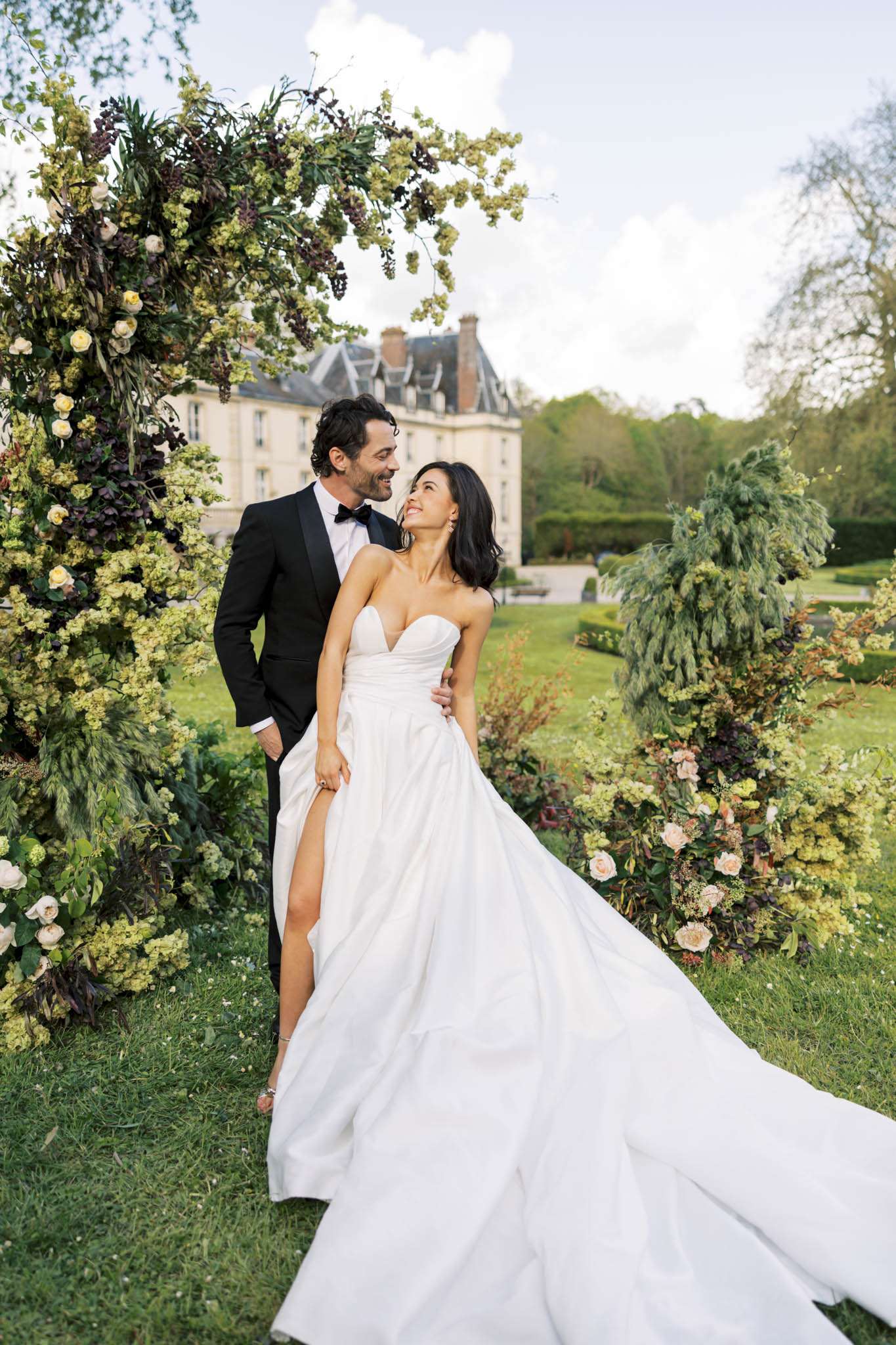 Couple smiles between garden-style floral arch columns with cream and blush roses before chateau