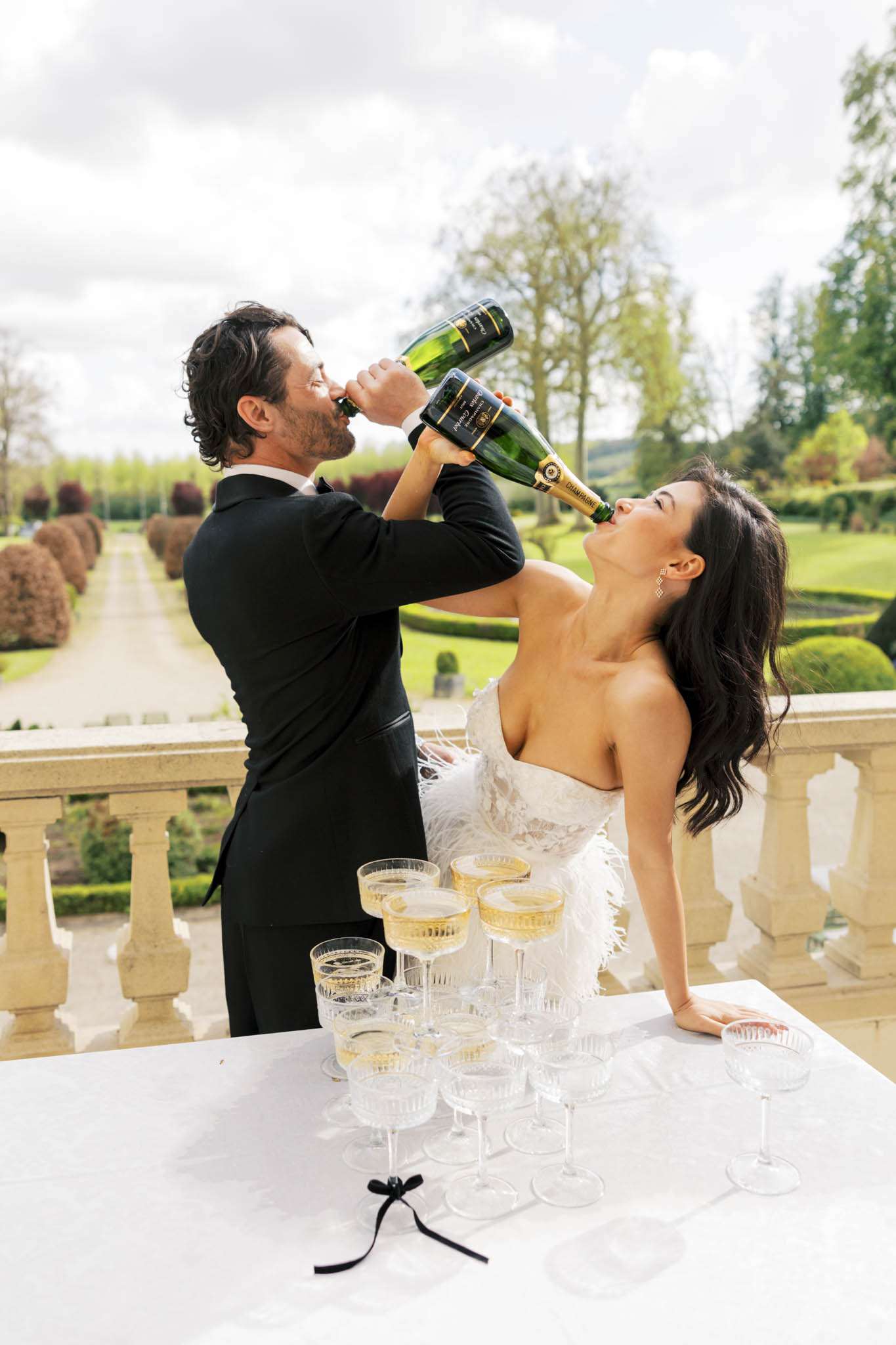 Couple drinking champagne from bottles beside crystal coupe tower on chateau terrace with formal gardens behind