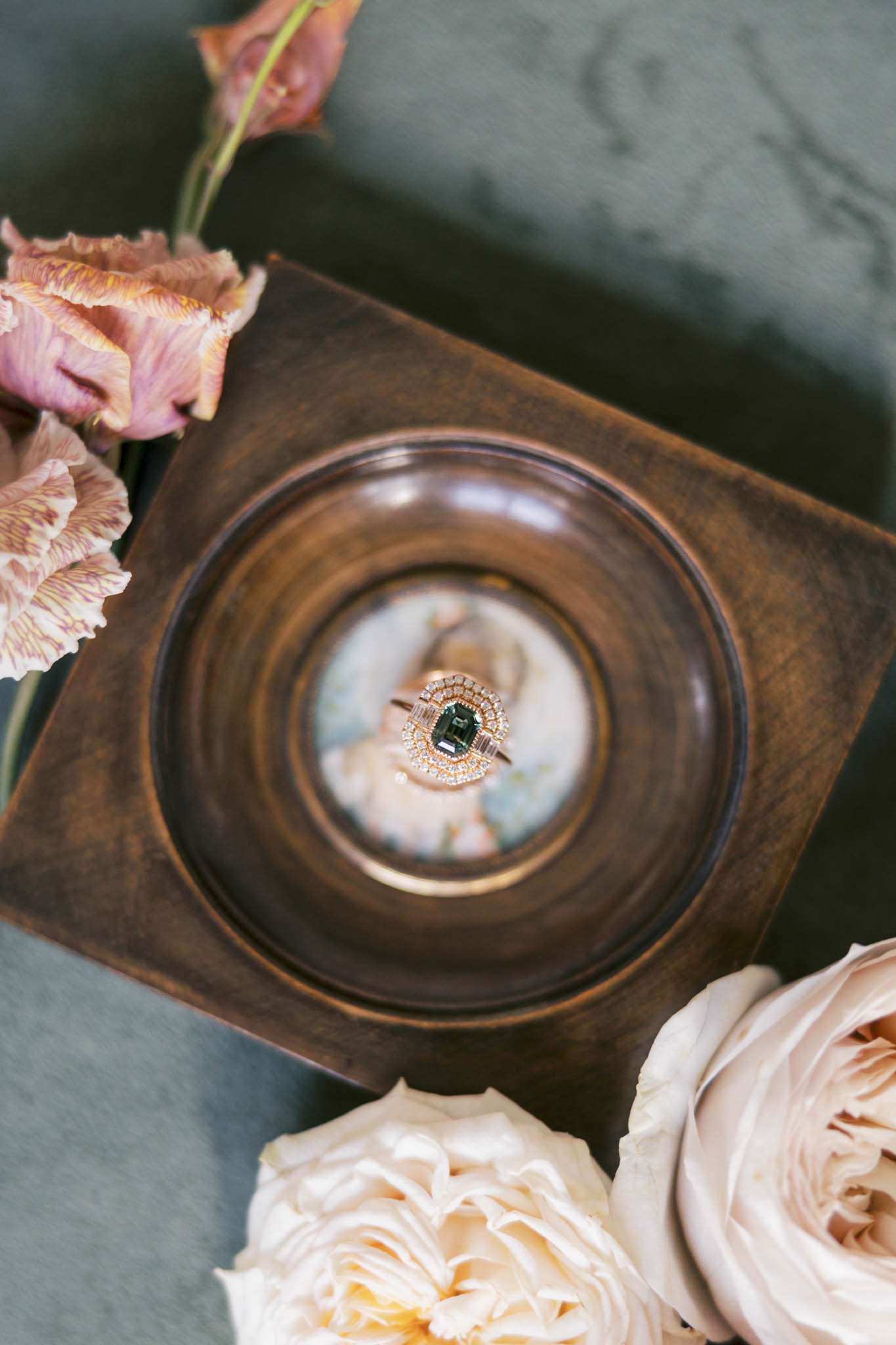 Engagement ring with teal-green oval stone in rose gold setting displayed in wooden dish with blush roses