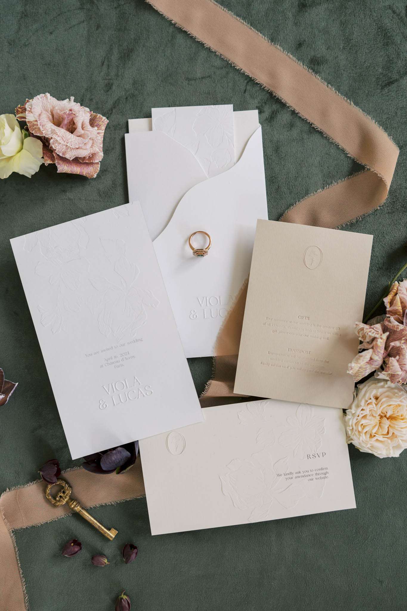 A flat lay detail shot of a wedding stationery suite for Viola and Lucas, arranged on a dark forest green velvet surface. The suite includes an ivory embossed invitation card, a matching envelope with a decorative wavy flap, a cream/sand-toned insert card with sections for gifts and transport information, and an ivory RSVP card — all featuring blind embossed figurative illustrations and an oval cameo motif. A gold engagement ring with a green center stone is placed at the center of the arrangement, accompanied by a vintage brass key, a loosely draped frayed-edge sand-colored silk ribbon, scattered mauve and cream lisianthus and garden rose blooms, and small fallen petals. The overall design palette is ivory, cream, and sand with a classic yet modern typographic style.