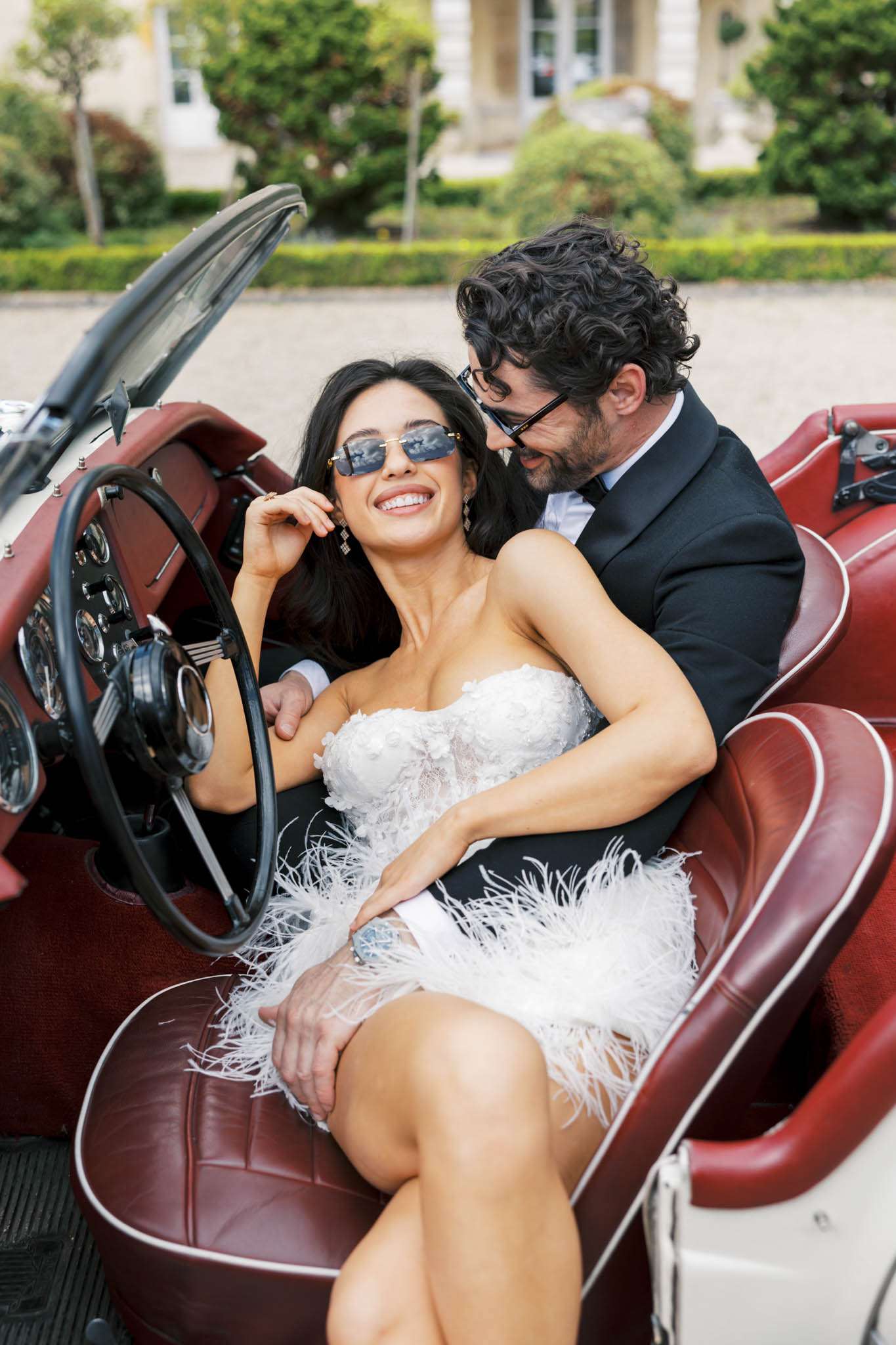 A couple portrait taken outdoors in front of what appears to be a French château, with the bride and groom seated together in a vintage convertible car with cream bodywork and deep burgundy red leather interior. The bride wears a short white strapless mini dress with 3D floral appliqué bodice and white feather trim hem, paired with small rectangular tinted sunglasses and drop earrings; she is laughing and leaning back against the groom. The groom wears a black tuxedo with a white dress shirt and bow tie, also wearing dark-framed sunglasses, and leans in toward the bride with his arm around her. The styling is modern and fashion-forward with a playful, editorial feel. The shot is a close-up portrait framed inside the car interior.