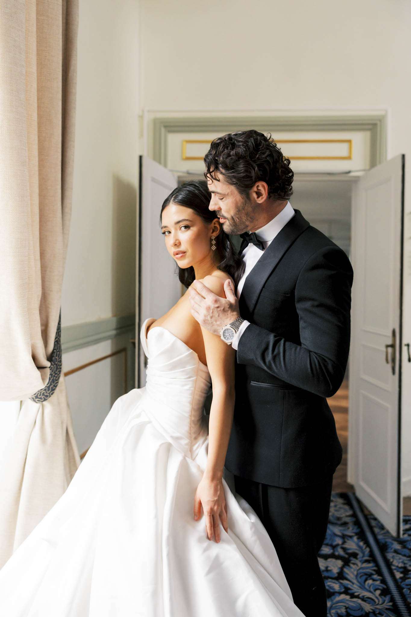 A couple portrait shot indoors in what appears to be a grand hotel or château corridor, with the groom standing behind the bride and resting his hands on her shoulders. The bride wears a white strapless ballgown with a structured bodice and full skirt, accessorized with drop earrings, her dark hair styled in a low updo. The groom wears a black tuxedo with a bow tie and a silver watch. The interior setting features white paneled walls with gold-trimmed molding, an ornate blue and navy patterned carpet, cream linen curtains with a blue-printed tieback, and white double doors visible in the background. The styling is classic and formal. Medium portrait composition, shot with soft natural window light from the left.
