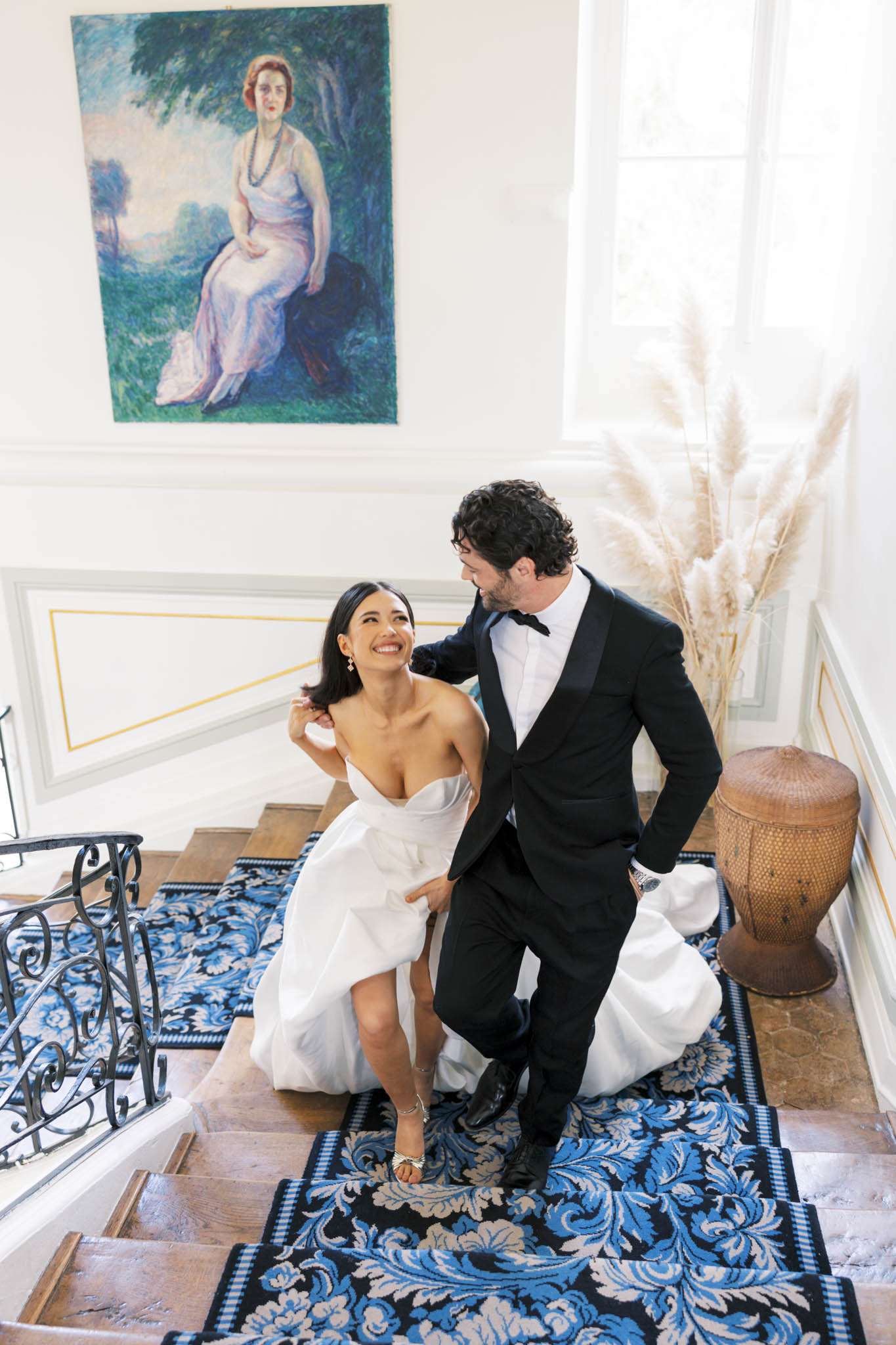 A couple portrait taken indoors on a grand staircase, shot from above at a slight angle. The bride wears a strapless white ballgown with a structured, ruffled skirt and strappy heeled sandals, and is smiling up at the groom while lifting her dress slightly as they ascend the stairs. The groom is dressed in a black tuxedo with satin lapels and a black bow tie. The staircase features a black, blue, and grey floral patterned runner and a wrought-iron railing. The interior space has white panelled walls with gold trim detailing, and is decorated with a large impressionist-style oil portrait of a woman in a pale pink dress hung on the wall, and a tall arrangement of dried pampas grass in a vase to the right. A woven decorative urn sits on the landing nearby, adding to the classic French interior setting.