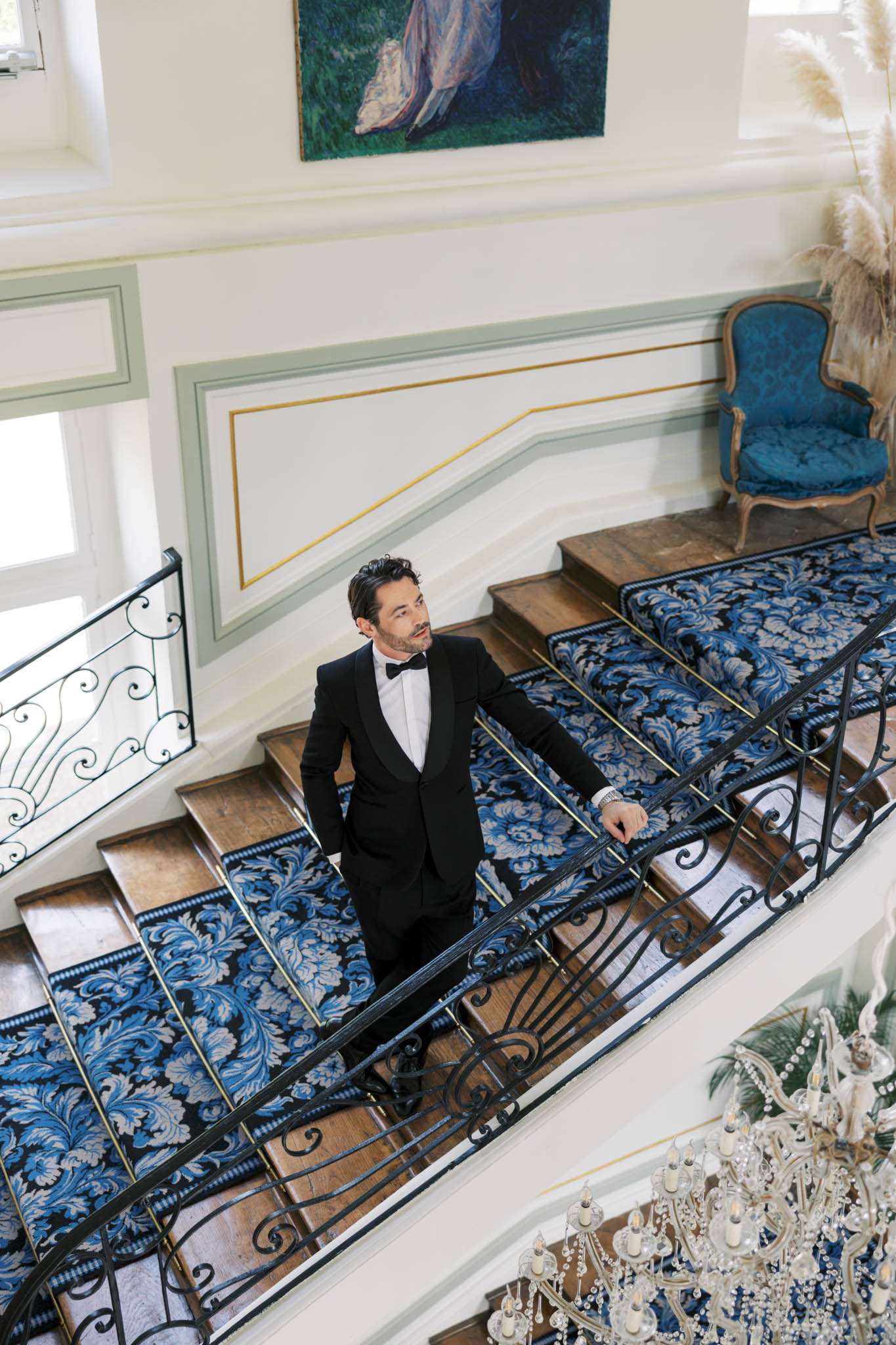 Overhead groom on grand staircase with damask carpet runner, blue velvet armchair, and crystal chandelier