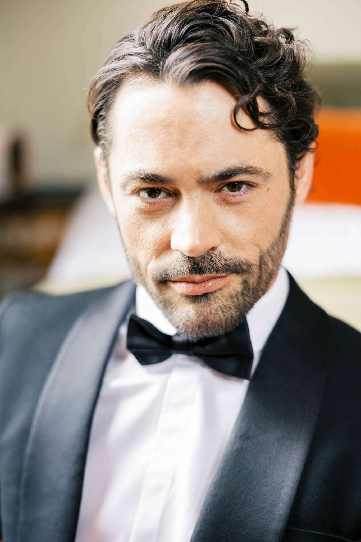 Close-up groom portrait in charcoal tuxedo with satin lapels black bow tie curly hair and short beard indoors