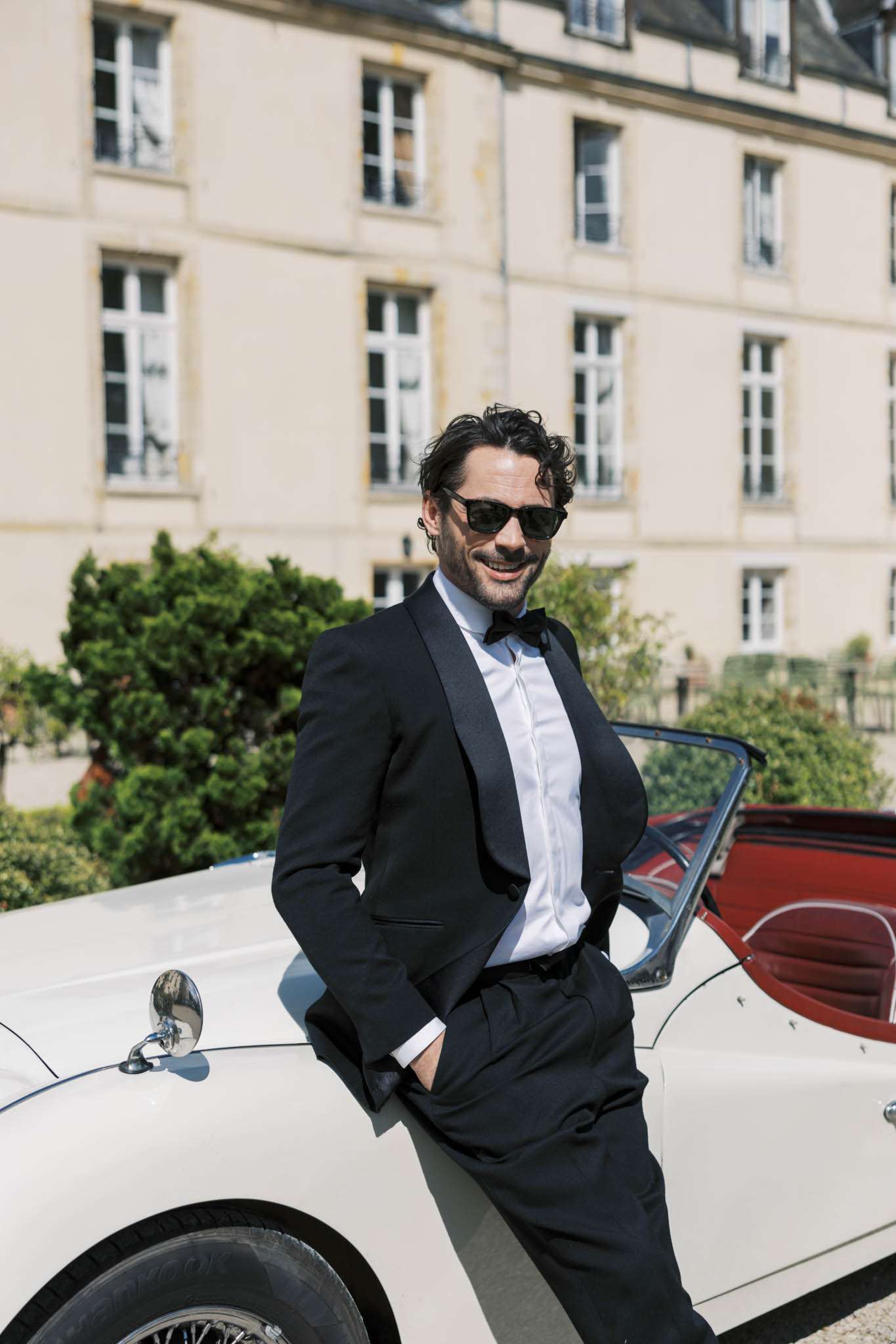 A groom portrait taken outdoors in front of a French château, with the building's cream stone facade and tall rectangular windows visible and softly out of focus in the background. The groom is leaning casually against a vintage cream convertible with red leather interior, hands in pockets, smiling and wearing dark sunglasses. He is dressed in a black tuxedo with shawl lapels, white dress shirt, and black bow tie, styled in a classic black-tie look. The shot is a mid-length portrait with natural daylight creating strong contrast.