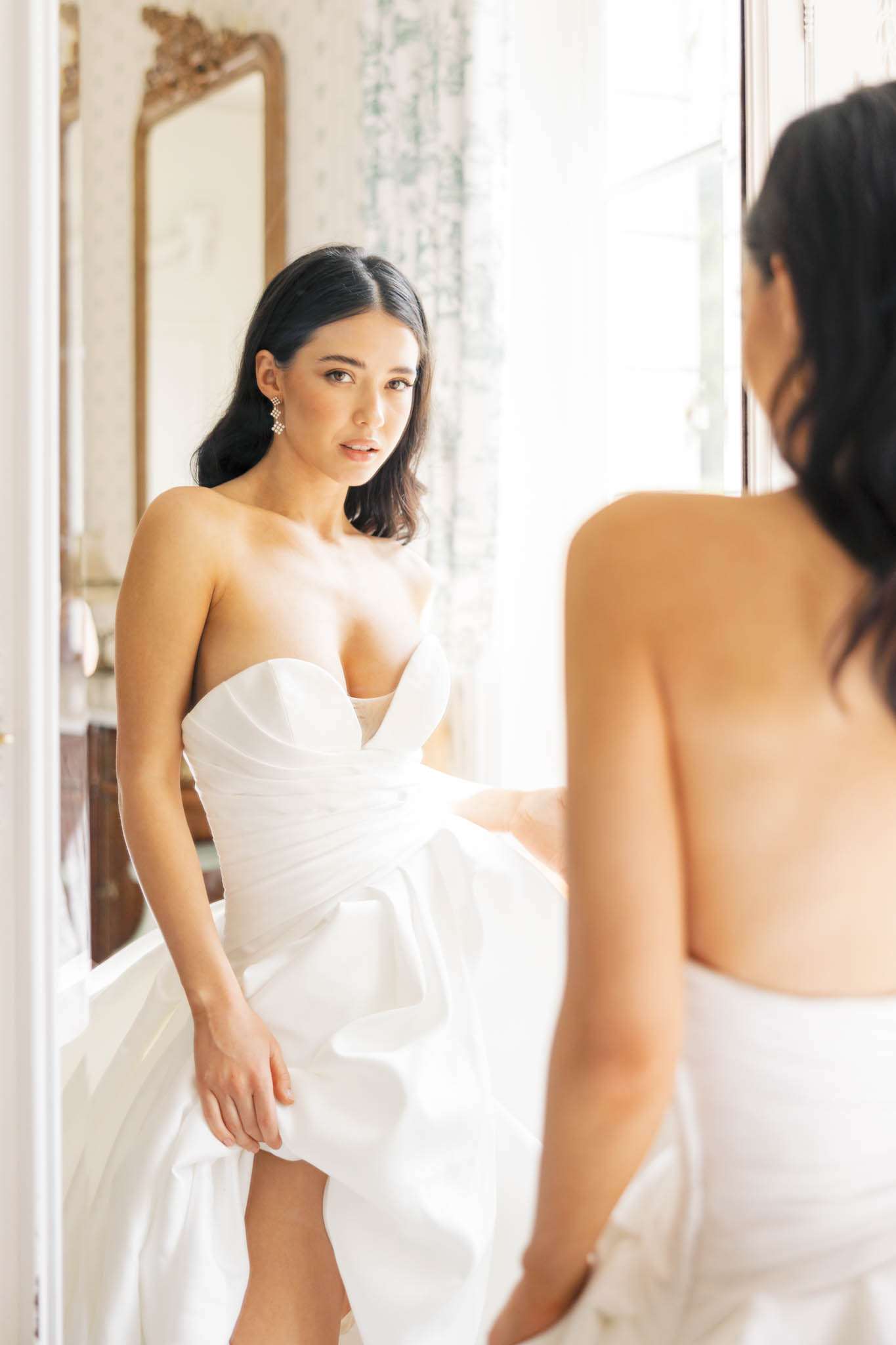 A bridal getting-ready portrait of a bride standing in front of a large ornate gold-framed mirror in an indoor room with decorative wallpaper and sheer white curtains. The bride wears a white strapless structured wedding dress with a deep sweetheart neckline and draped, voluminous skirt detailing; she accessorizes with geometric drop earrings and her dark hair is worn down in loose waves. The composition is a medium portrait shot captured from slightly behind and to the side, showing both the bride directly and her reflection in the mirror simultaneously. The room features classic French interior styling with gilt-framed furniture visible in the background, and the overall aesthetic is modern and clean.