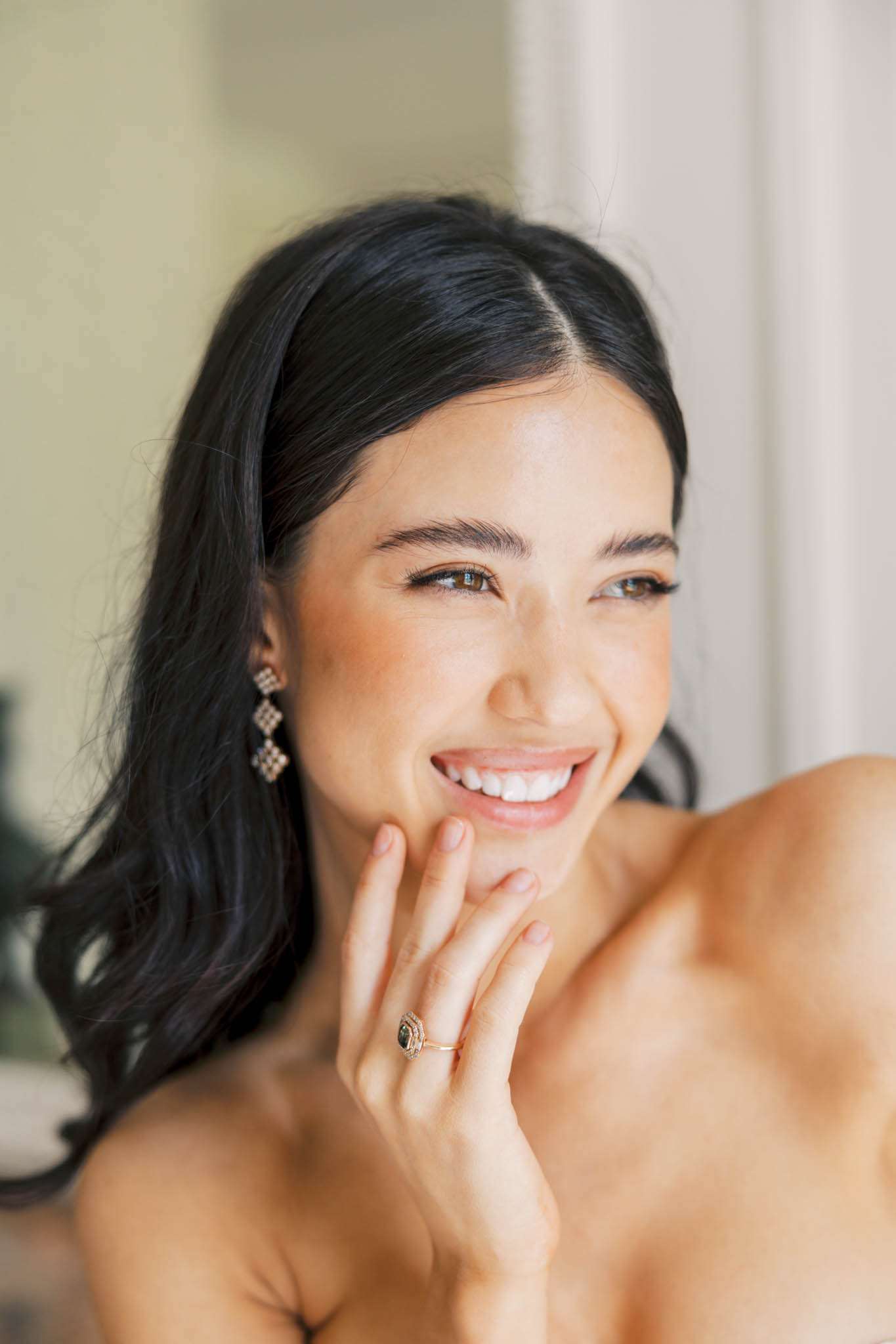 Close-up bridal portrait with dark hair, geometric gold drop earrings, and green stone engagement ring in gold halo setting