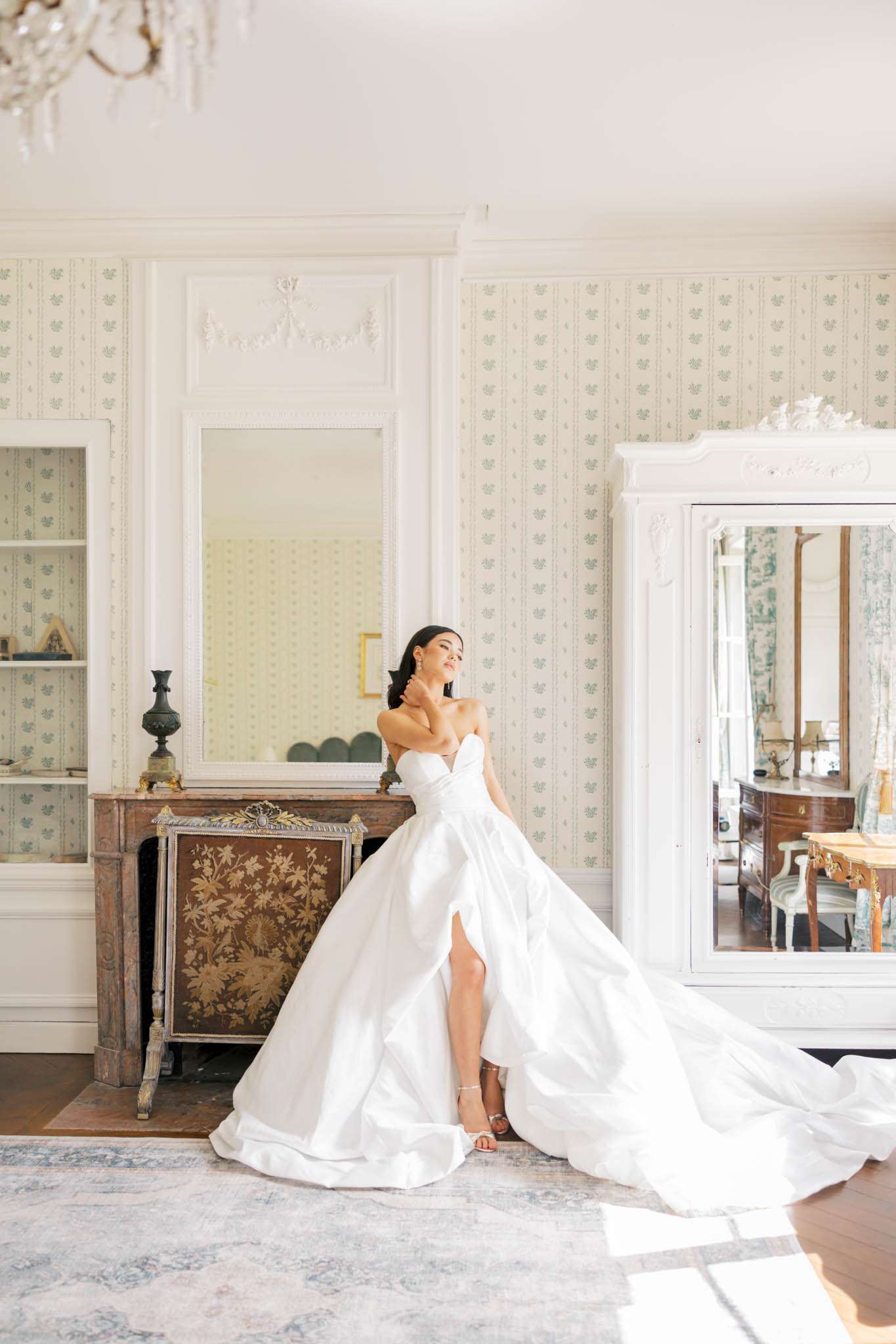 A bridal portrait taken indoors in what appears to be a château getting-ready room. The bride, a dark-haired woman, poses alone in a white strapless ballgown with a sweetheart neckline, a high front slit, and an extended cathedral-length train that pools across the floor; she wears strappy silver heels and drop earrings, with one hand raised to her neck. The room features pale green and white floral wallpaper, white wood-panelled boiserie with ornate plasterwork, an antique marble and gilt fireplace with a decorative gold and brown firescreeen, a large wall mirror above the mantle, and a white-painted armoire with mirrored doors to the right. A crystal chandelier is partially visible at the top of the frame, and a faded blue and grey area rug anchors the space, giving the room a classic French interior aesthetic. Full-length portrait composition shot in natural light streaming from one side.