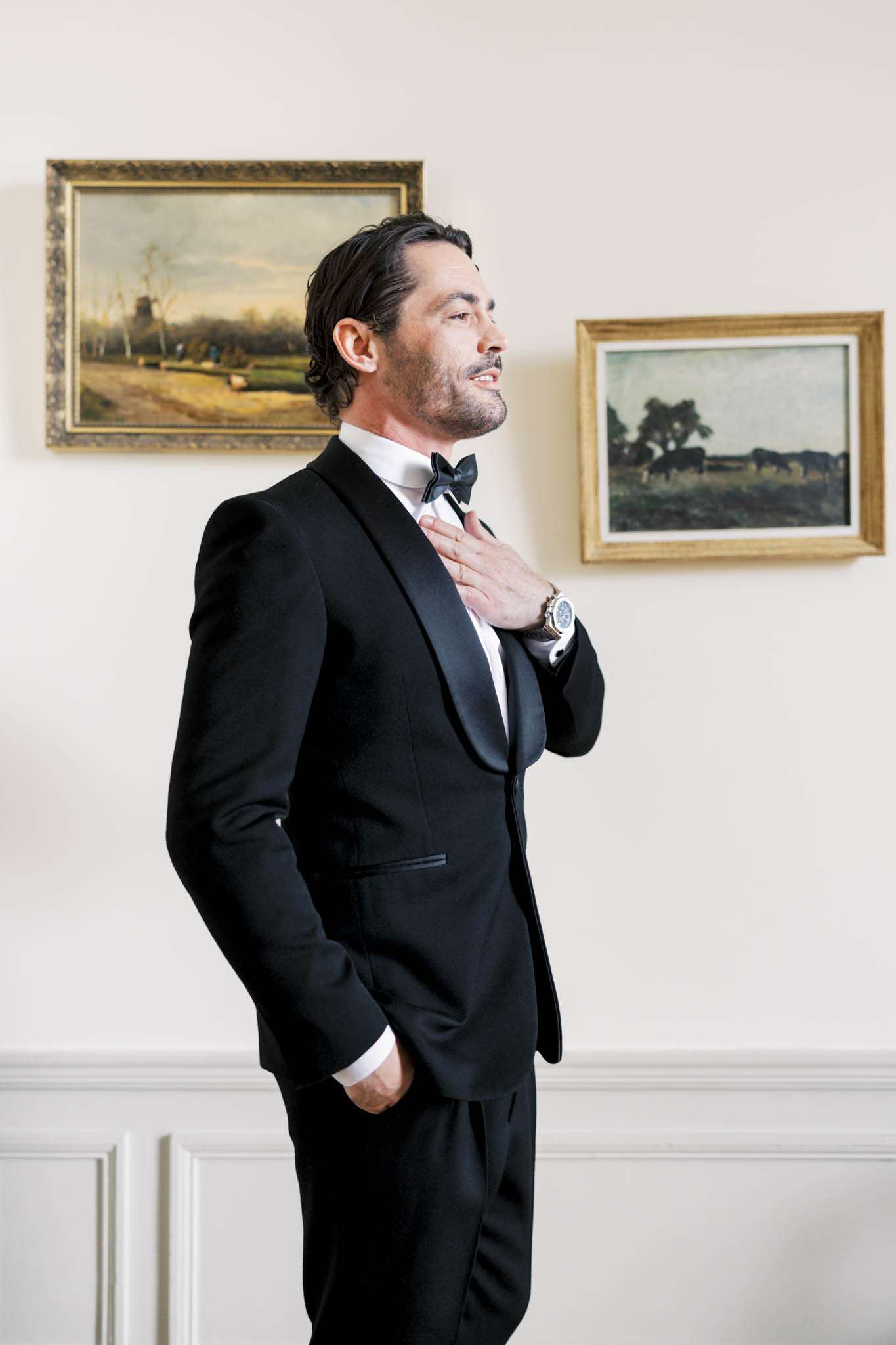 Groom adjusting black bow tie wearing tuxedo with satin lapels in chateau interior with oil paintings