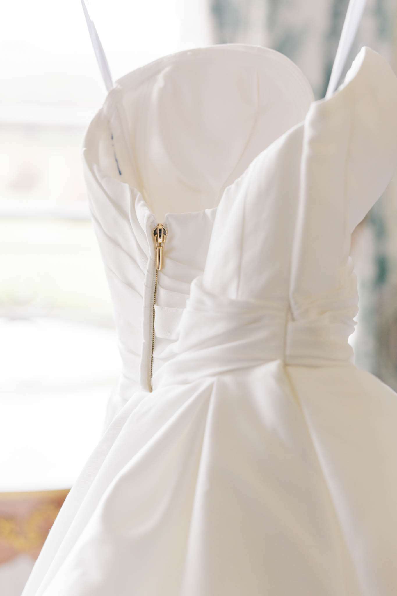 White wedding dress back detail showing gold zipper and draped satin bodice hanging near window light