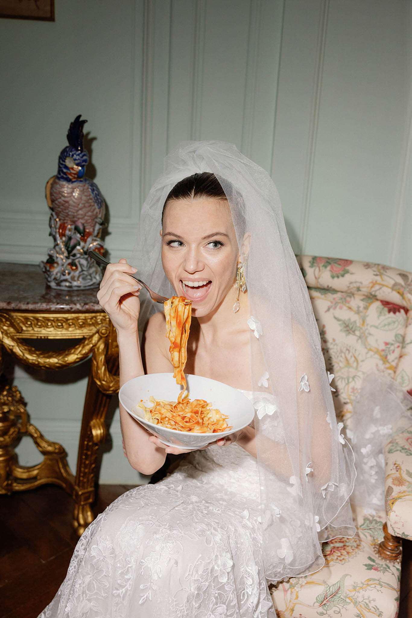 Bride in strapless ivory lace gown laughing while eating pasta on floral sofa inside chateau salon