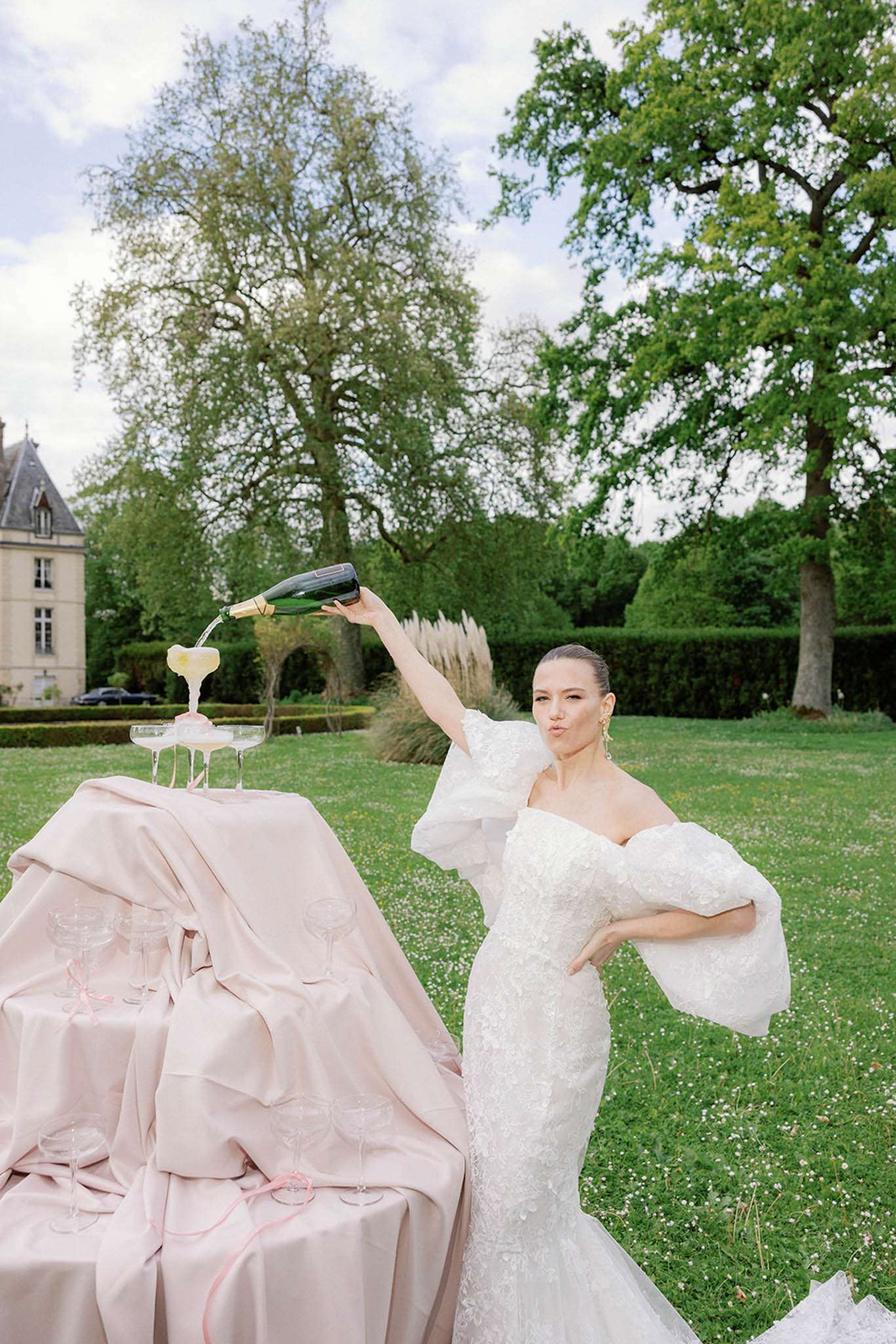 Bride in lace puff-sleeve gown pouring champagne into a coupe tower on blush satin at a French chateau
