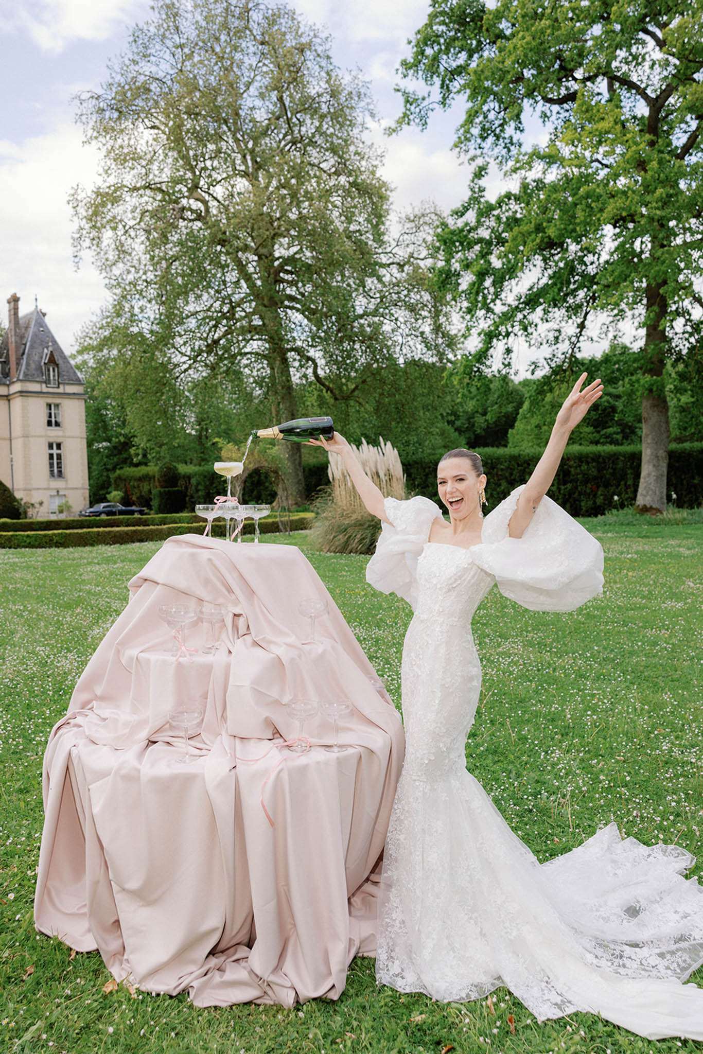 A bride pours champagne into a tower of coupe glasses outdoors on the manicured grounds of a French château, raising one arm in celebration with a wide smile. She wears a fitted white lace mermaid gown with dramatic oversized off-the-shoulder puff sleeves and a long lace train spread across the lawn. The champagne tower is set on a round table draped in a blush pink silk linen, with additional coupe glasses arranged on the table. A classic French château with slate-roofed turrets is visible in the background to the left. The shot is a full-length portrait taken at a slight distance, capturing both the bride and the full table setup in a candid, celebratory moment.